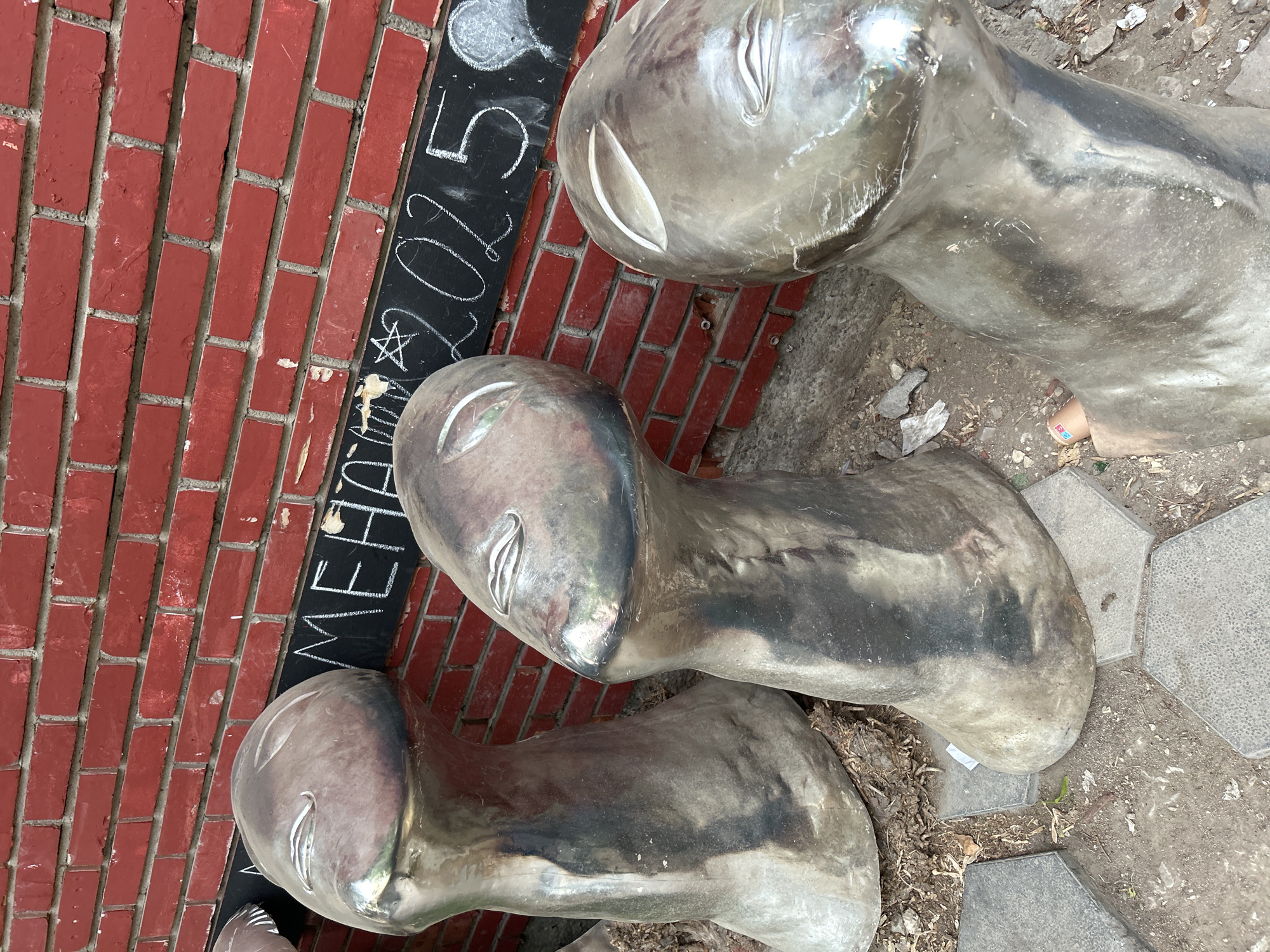 Three silver-colored, abstract, sculptural heads with simplified facial features, resembling stylized human or alien forms, are positioned at an angle next to a red brick wall on which the words "MEHAK 2015" are written in white chalk. The heads appear to be resting on a rough surface of dirt, concrete and some dry leaves. The location is most likely in the city of Plovdiv, Bulgaria.