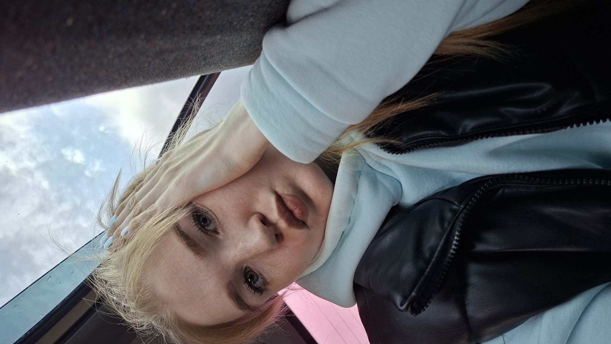 A person is lying down, seemingly inside a vehicle, with their hand covering part of their forehead. The person has fair skin, light-colored hair, and is wearing a light blue hooded sweatshirt and a black vest. The background shows a cloudy sky visible through the car window. The location is Chaikovsky, Russia.