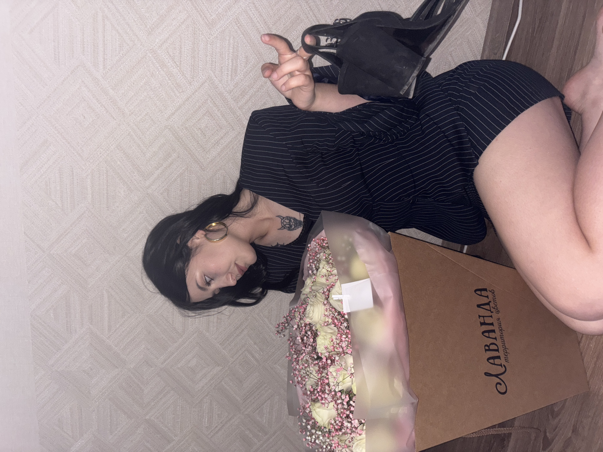 A young woman with dark hair and gold hoop earrings kneels on a wooden floor next to a large bouquet of white roses and pink baby's breath in a brown cardboard box. The box has the inscription "ЛАВАНДА территория цветов" in black lettering. She is wearing a pinstriped black blazer over a dark top and is holding a black high-heeled shoe in her right hand. A tattoo of a horned, grinning face is visible on her chest. The background is a wall with a geometric pattern. The scene appears to be indoors, possibly in an apartment, in the city of Chaikovsky, Russia.