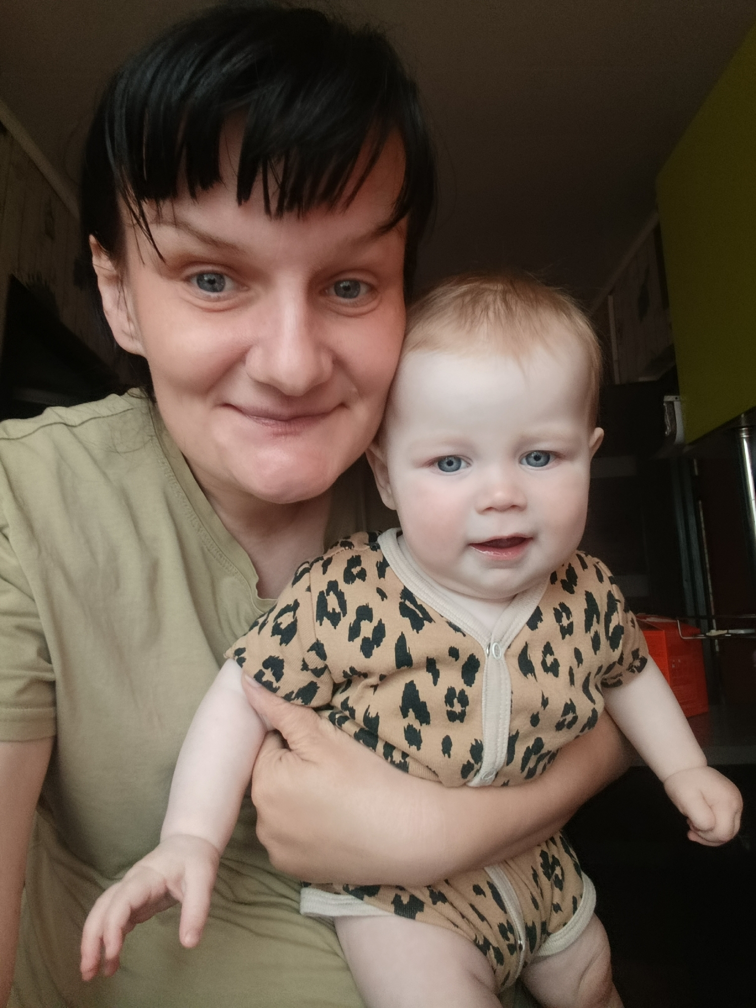 A close-up, informal portrait features a woman holding a baby indoors. The woman, positioned on the left, has dark, short hair with bangs and blue eyes, and is smiling gently at the camera. She wears a plain, light olive green t-shirt. The baby, held securely in her arms and located on the right, has light blonde or reddish hair and bright blue eyes, looking towards the camera with a curious, slightly open-mouthed expression. The baby is dressed in a tan romper or onesie with a distinctive black leopard print pattern and a front zipper. The background is dimly lit and indistinct, suggesting an indoor setting with subtle wall textures and a hint of a light green surface or object on the far right. Both subjects appear engaged with the camera, conveying a warm and affectionate interaction. No visible text is present.