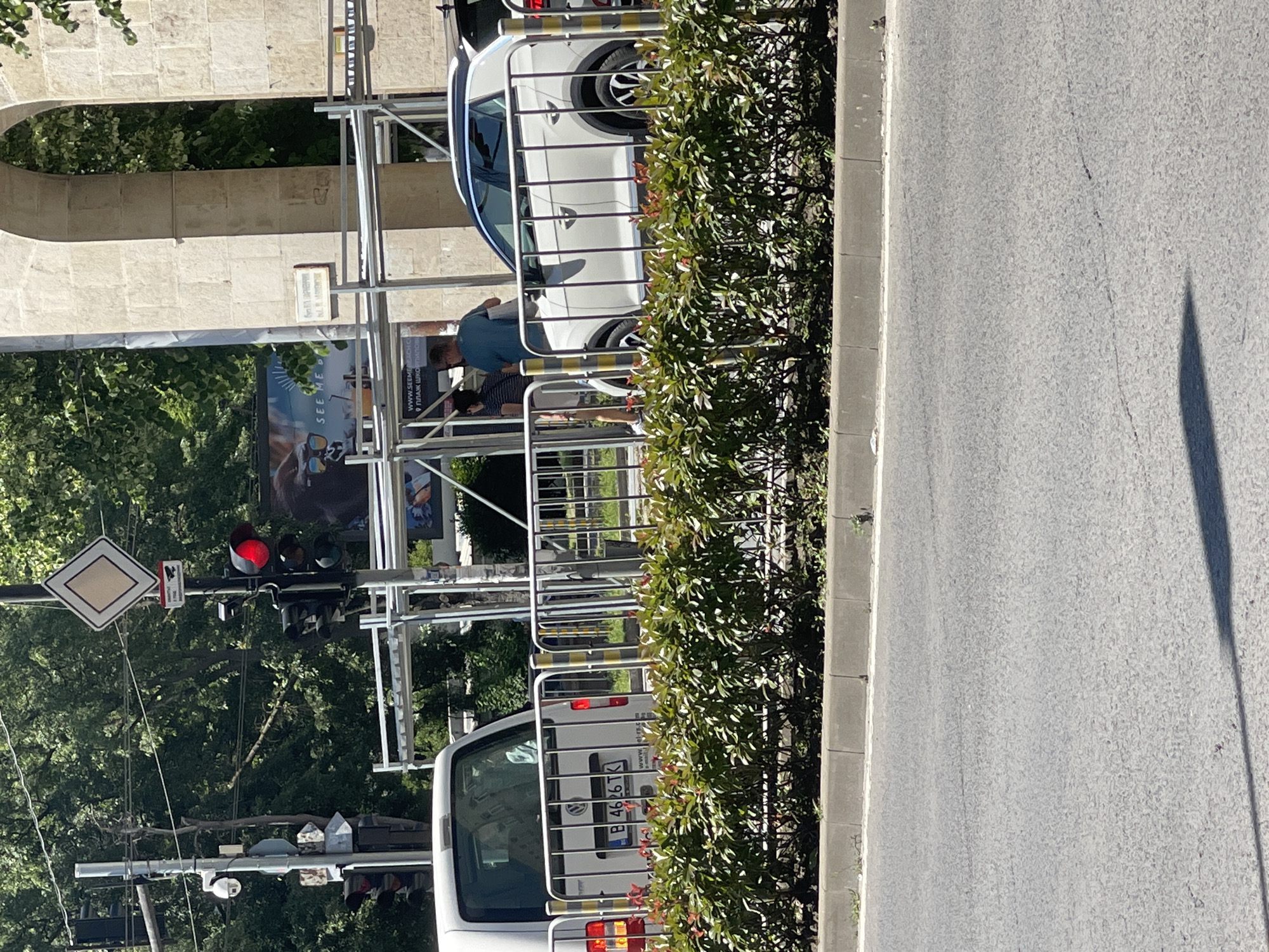 The image shows a street scene in Varna, Bulgaria, with a traffic light displaying red. A white Volkswagen van and a white Volkswagen T-Roc are parked on the side of the road, behind a metal fence and a row of green bushes. Two individuals, a woman and a man, are standing near the Volkswagen T-Roc, conversing. Scaffolding is erected behind them, suggesting ongoing construction or maintenance work. In the background, a large stone archway with ornate carvings is visible, as well as trees and a billboard. The street is paved, and a sidewalk is present with a concrete curb. The sun is shining, casting shadows on the road.