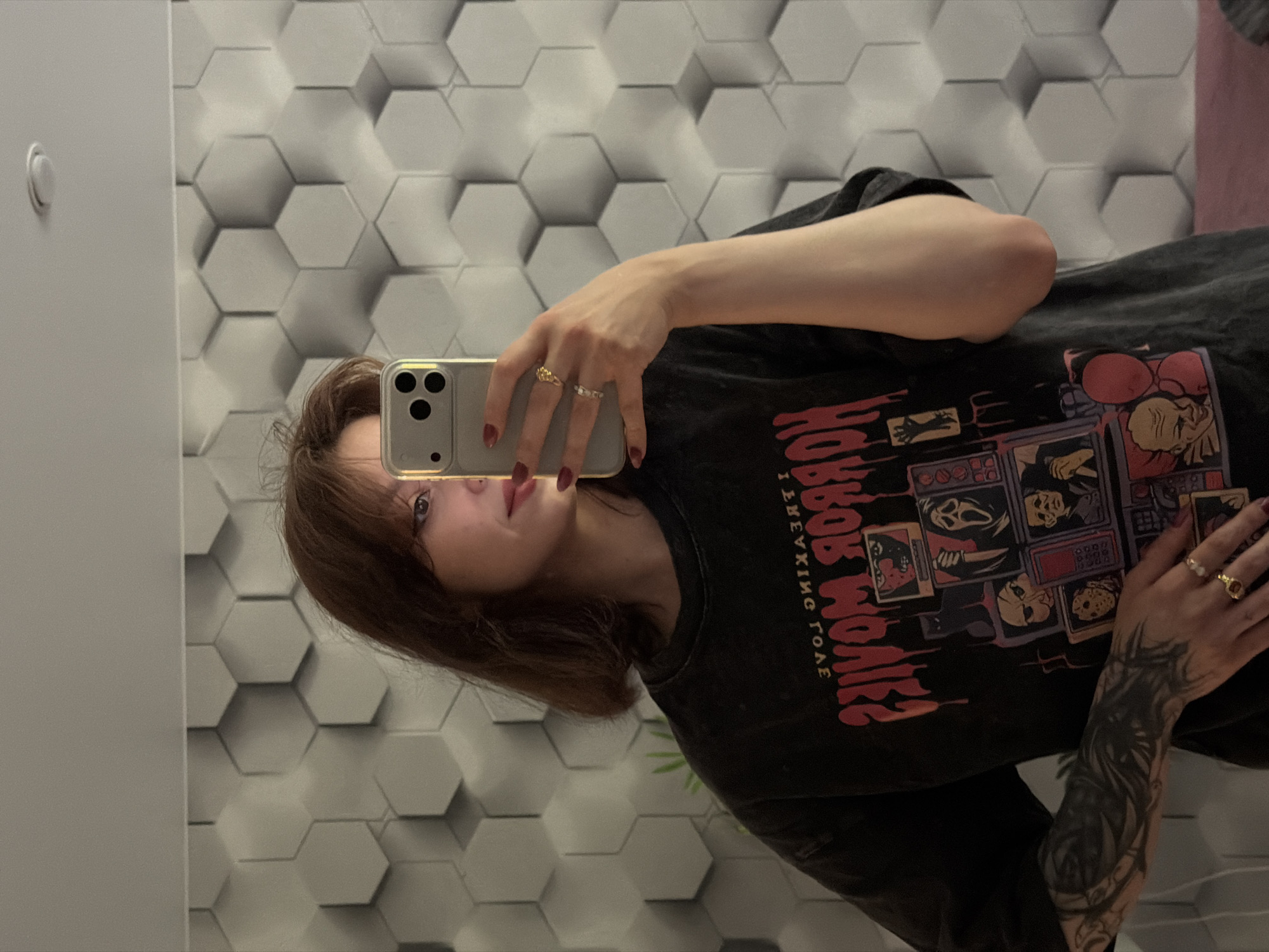 A young woman with shoulder-length brown hair and fair skin is taking a mirror selfie in what appears to be a residential interior. She is wearing a dark-colored t-shirt with a graphic print depicting various horror movie characters, including Freddy Krueger, Jason Voorhees, and Ghostface, arranged in a collage-like fashion. The text on the shirt reads "TERROR MOVIES" in a blood-like font, although it is partially reversed due to the mirror. She has dark red nail polish on her fingernails and is wearing several rings on her fingers. Her left arm, visible in the foreground, is adorned with an intricate black tattoo that extends from her wrist up her forearm. The background consists of a textured hexagonal wall pattern in varying shades of grey and white, and a ceiling light fixture is visible. The lighting is soft, creating a slightly dim ambiance. The scene is captured from the chest up, with the woman holding a silver smartphone to take the photo.