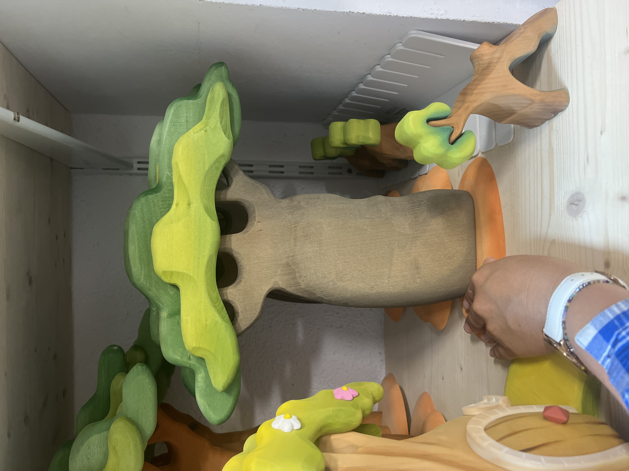 A close-up, eye-level shot shows a collection of wooden toy trees and a hand interacting with one of the toys. The toys are painted in shades of green and brown, with some featuring small white and pink flowers. The main focus is a large tree with a brown trunk and layered green and yellow foliage. To the left, a portion of another toy tree, resembling a house with a circular door, is visible. A human hand, adorned with a white watch and a colorful beaded bracelet, is gently touching the base of the larger tree. The toys are displayed on a light-colored wooden shelf, which appears to be part of a shelving unit. In the background, a white fence-like object and a wall are visible. The lighting is soft and diffused, illuminating the toys and the hand. The scene is indoors, likely in a playroom or a retail setting.