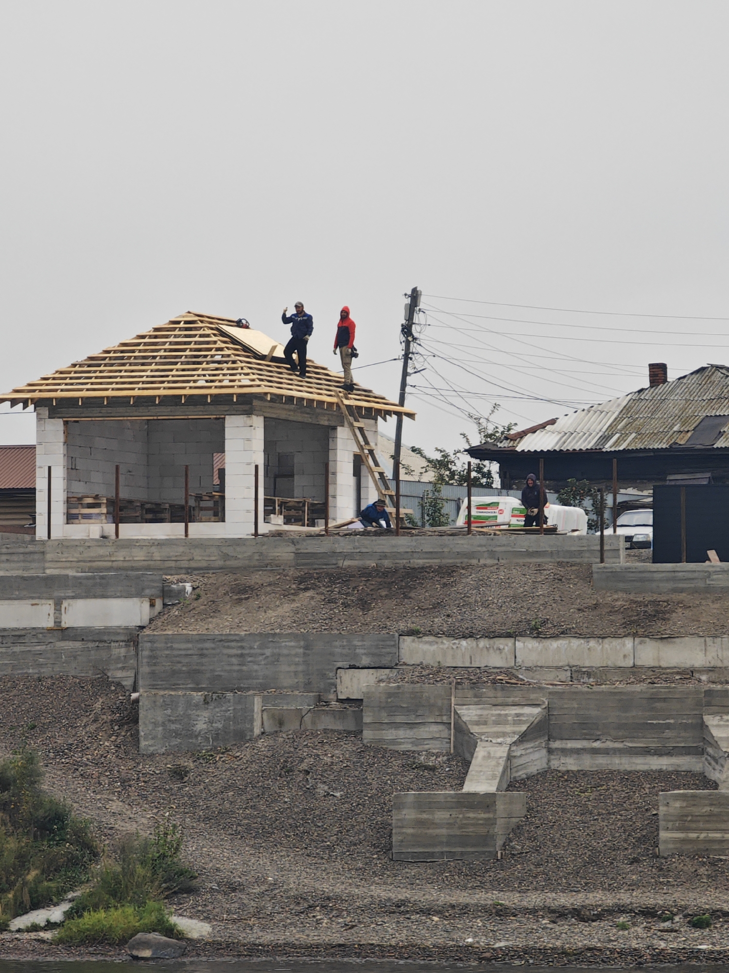 In Ермолаево, Russia, a construction site is depicted. A building with a wooden roof frame is under construction, with several individuals present. Two men are on the roof, one gesturing. Another man is near a ladder, and a fifth person is on the ground near the building's foundation. The structure is made of white concrete blocks. In the background, an older, weathered building with a corrugated metal roof stands next to a utility pole with electrical wires. Construction materials, including pallets of white bags, are visible near the older building. The foreground shows a tiered embankment made of concrete and gravel, with vegetation growing on the lower slopes. A body of water is at the bottom of the frame, with a rock in the foreground. The sky is overcast and a light grey.