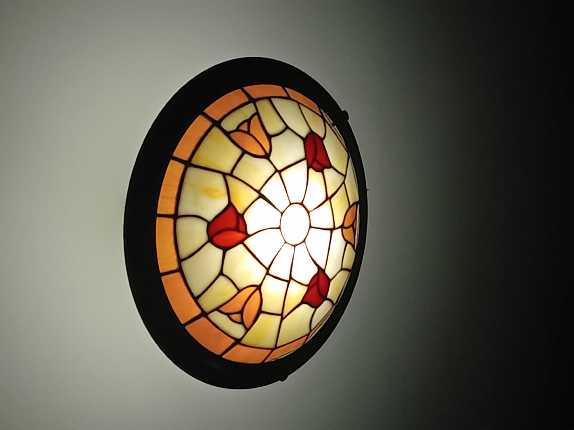 A close-up shot depicts a round, stained-glass ceiling light fixture. The fixture has a black circular frame, and the stained-glass design inside features a floral pattern. The glass is predominantly white with yellow undertones, outlined by a network of black lines. Within the white and yellow glass, there are stylized floral motifs in shades of red and orange. The light source is located at the center of the fixture, emitting a bright glow that illuminates the surrounding glass. The light is mounted on a surface that transitions from a light grey on the left to a dark grey/black on the right, suggesting a gradient or shadow effect. The location is in the city of Unknown, Unknown.