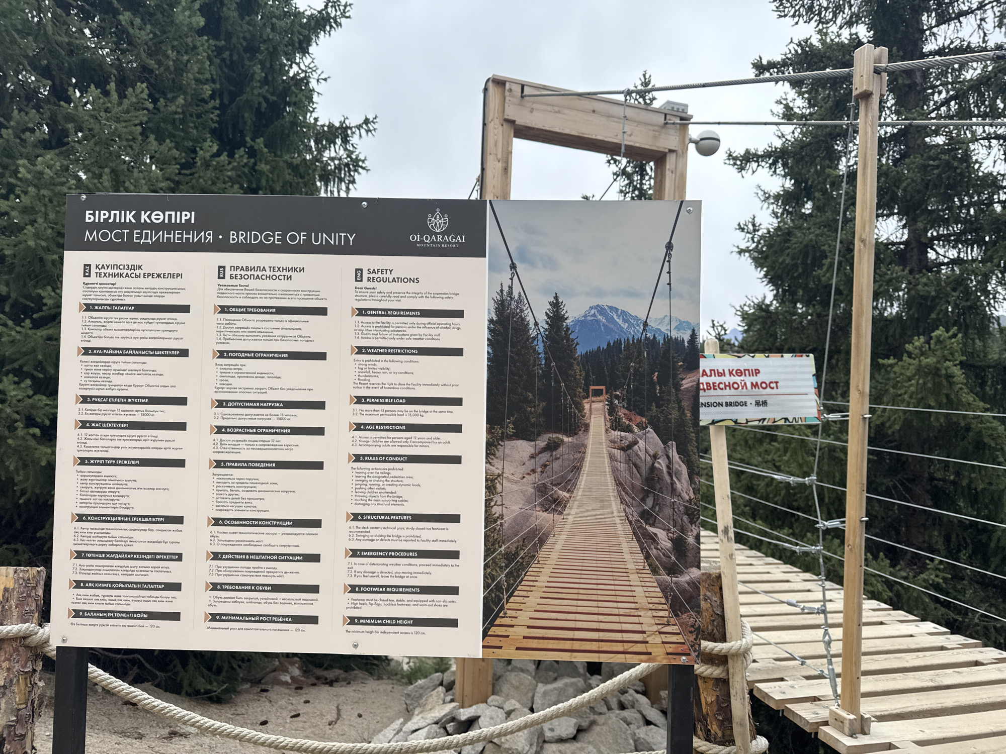 The image shows a sign for the "Bridge of Unity" at the Oi-Qaragai Mountain Resort, located in the city of Almaty, Kazakhstan. The sign is multi-lingual, featuring text in Kazakh, Russian, and English, and displays safety regulations and information about the bridge. The background of the sign features a photograph of the wooden suspension bridge itself, spanning across a rocky gorge with a majestic mountain in the distance. The environment appears to be a mountainous, forested area, with lush green trees surrounding the structure. The sign is positioned at the entrance to the bridge, and part of the bridge's wooden walkway and supporting cables are visible in the foreground, along with some rocks and rope. The sky is overcast.
