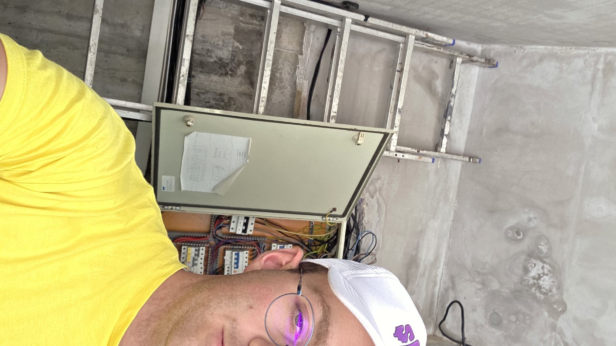 A medium shot captures a person wearing a bright yellow t-shirt and a white baseball cap with a purple logo. The person is also wearing round glasses that reflect a purple hue. They are positioned in the foreground, partially obscuring a gray electrical panel that is open, revealing a complex arrangement of wires and circuit breakers. Behind the electrical panel, a silver ladder leans against a textured gray wall. The ceiling appears unfinished and textured, with visible water stains. The scene suggests a construction or renovation environment.