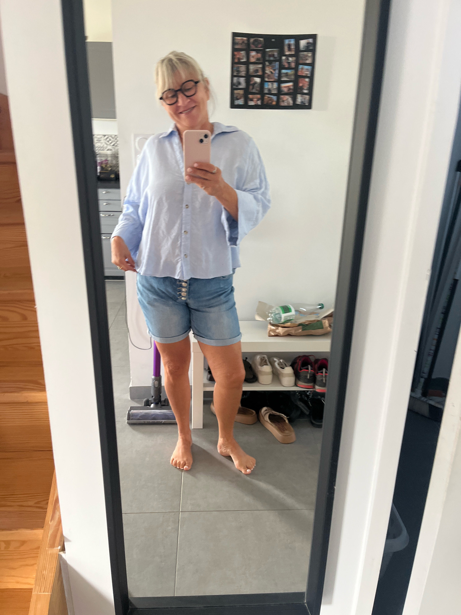 A woman is standing in front of a full-length mirror, taking a selfie. She is wearing a light blue oversized button-up shirt and denim shorts. She has blonde hair and is wearing round glasses. In the reflection, a hallway is visible with a wooden staircase on the left, a shoe rack filled with various shoes on the right, and a wall with a framed collage of photos. A vacuum cleaner is partially visible on the floor. The room appears to be a home interior. The image was taken in Chambœuf, France.