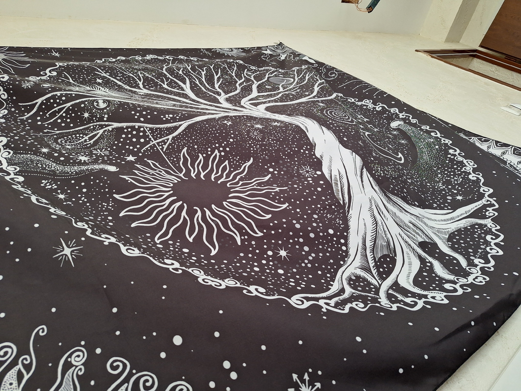 A black tapestry with a white celestial design is draped across a surface, likely a bed or the floor. The design features a tree with roots and branches encircled by a dotted border and a sun symbol. There are stars and other celestial motifs scattered throughout the design. The background behind the tapestry is a textured, off-white wall. An open doorway or hatch with a wooden frame is visible in the upper right corner. The image doesn't provide any information that can determine if the image was taken in Panagyurishte, Bulgaria.