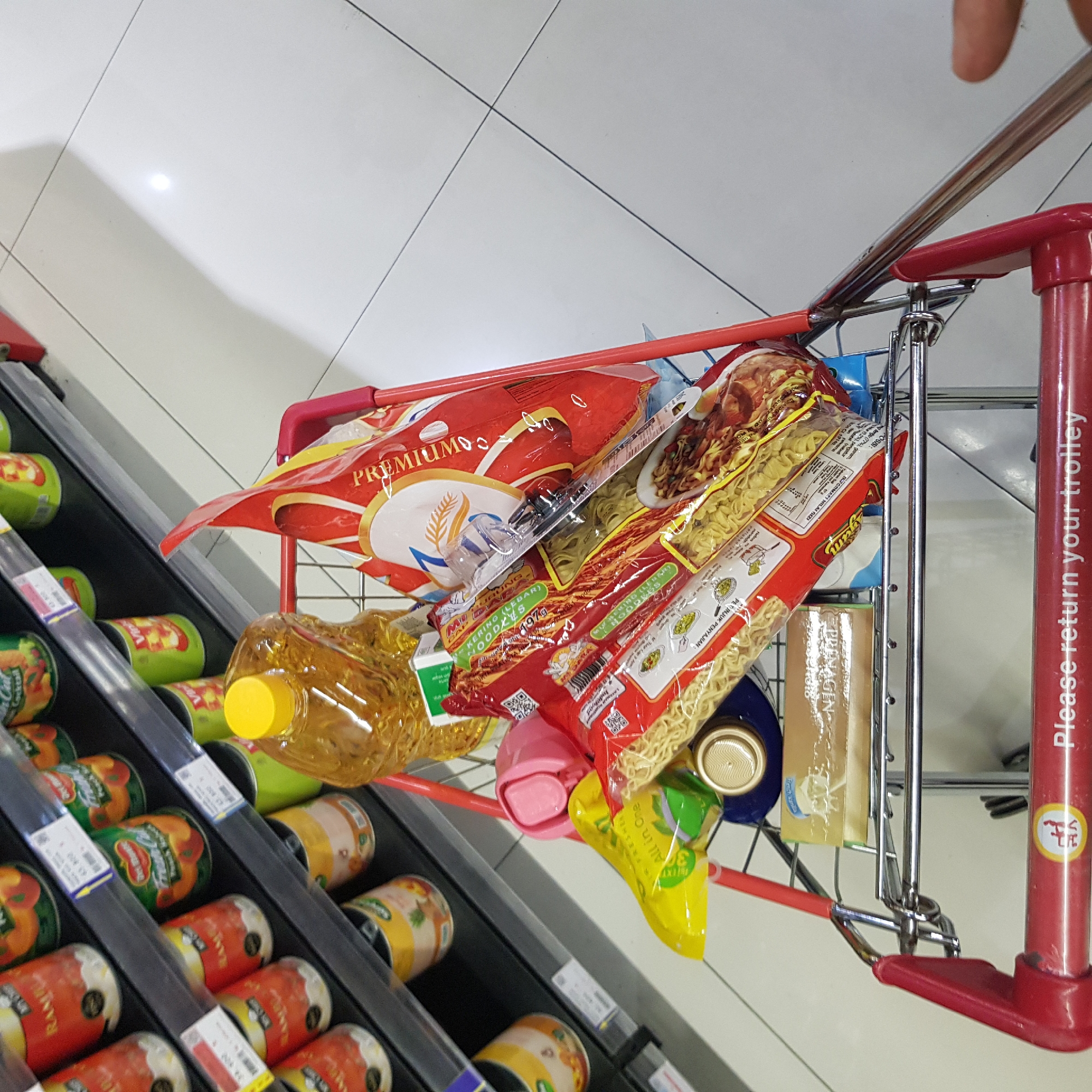 The image shows a shopping cart filled with groceries. The cart is red and silver, with a red handle and a silver wire basket. The groceries include several bags of instant noodles, a bottle of cooking oil, a yellow pouch, a pink object, and a box of what appears to be crackers or biscuits. The shelves behind the cart are stocked with cans and jars of food items, many of which are orange or green. The floor is tiled with white squares. The scene appears to be inside a supermarket. The text "Please return your trolley" is visible on the handle of the shopping cart. The context provided for Banda Aceh, Indonesia suggests that this image was taken in a supermarket in that city.