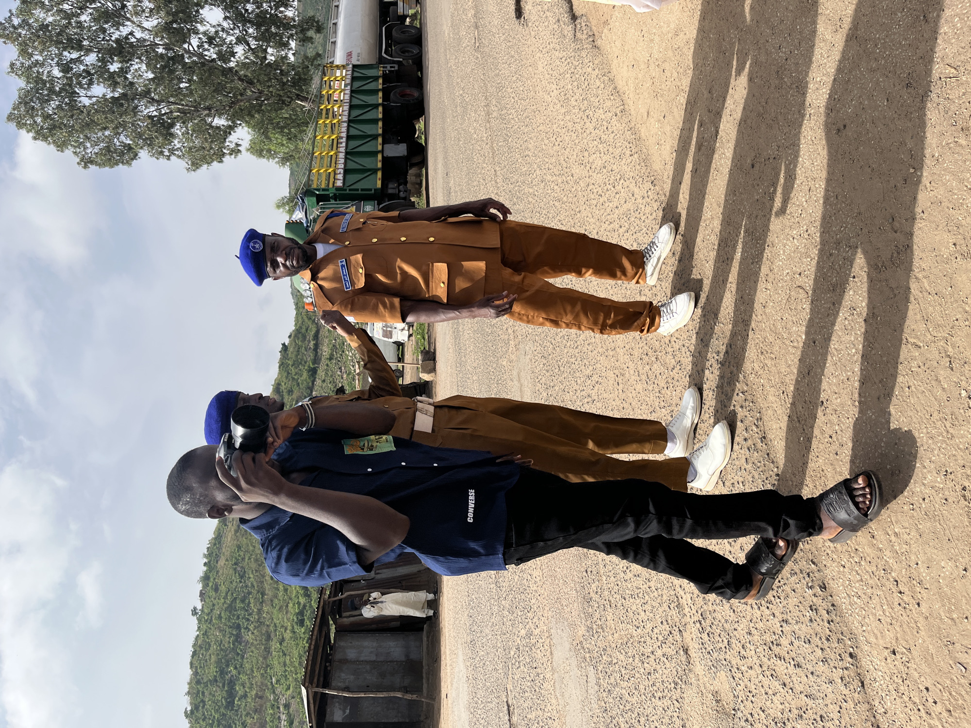 The image depicts three people on a road, possibly in the vicinity of Jos, Nigeria. Two individuals are wearing matching brown uniforms with blue berets and white sneakers. The third person, who is taking a photograph, is wearing a dark blue shirt with the word "CONVERSE" visible on the back, black pants, and black sandals. They are holding a black camera with a large lens. A large truck is parked nearby. The background shows a landscape with hills and trees under a cloudy sky. The setting appears to be a rural or semi-rural roadside.