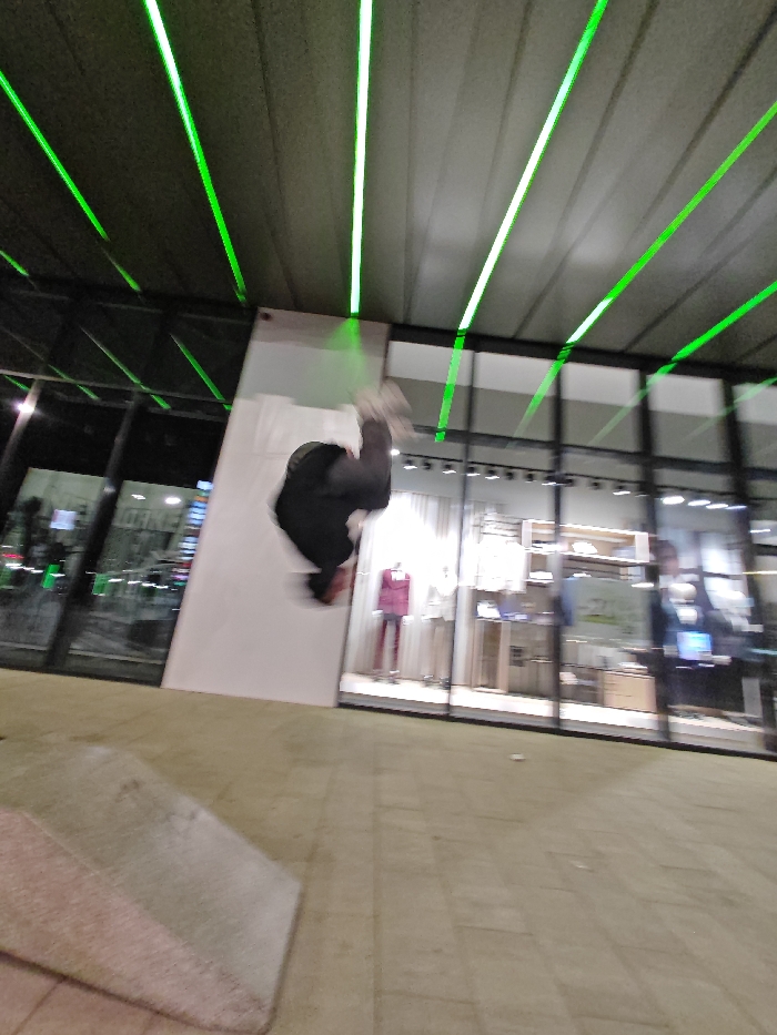 The image depicts an outdoor urban environment, likely at night, in Shumen, Bulgaria. A heavily motion-blurred individual, wearing dark clothing and light-colored footwear, is suspended mid-air against a prominent white, angled architectural panel, indicative of an acrobatic maneuver. The ground consists of light-colored, rectangular paving stones, with a low, light-toned concrete ramp visible in the immediate foreground on the left. To the left of the white panel, a dark, reflective glass facade of a building is visible, reflecting external lights. To the right of the panel, a retail storefront features large glass windows displaying mannequins in suits (one maroon, one light-toned) and other merchandise, with an illuminated interior and a visible "20%" discount sign. Overhead, a ceiling structure is illuminated by multiple parallel, bright green LED strip lights that stretch into the background.