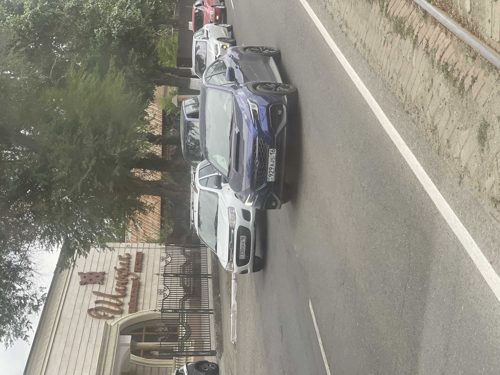 A dark blue Subaru WRX with license plate KZ 929 AJS 16 is in the foreground, driving on a paved road in Oskemen, Kazakhstan. To its left is a white car with license plate KZ 330 DPA 16. In the background, a building with the Cyrillic script "Шанграй" above a smaller inscription "МЕЙРАМХАНА РЕСТОРАН" is visible. The building is adorned with ornate architectural details and an iron fence. Several mature trees line the street, with lush green foliage. Other vehicles, including a silver sedan and a red sedan, are also present in the traffic flow. The road surface shows some signs of wear and cracking. The sky appears overcast.