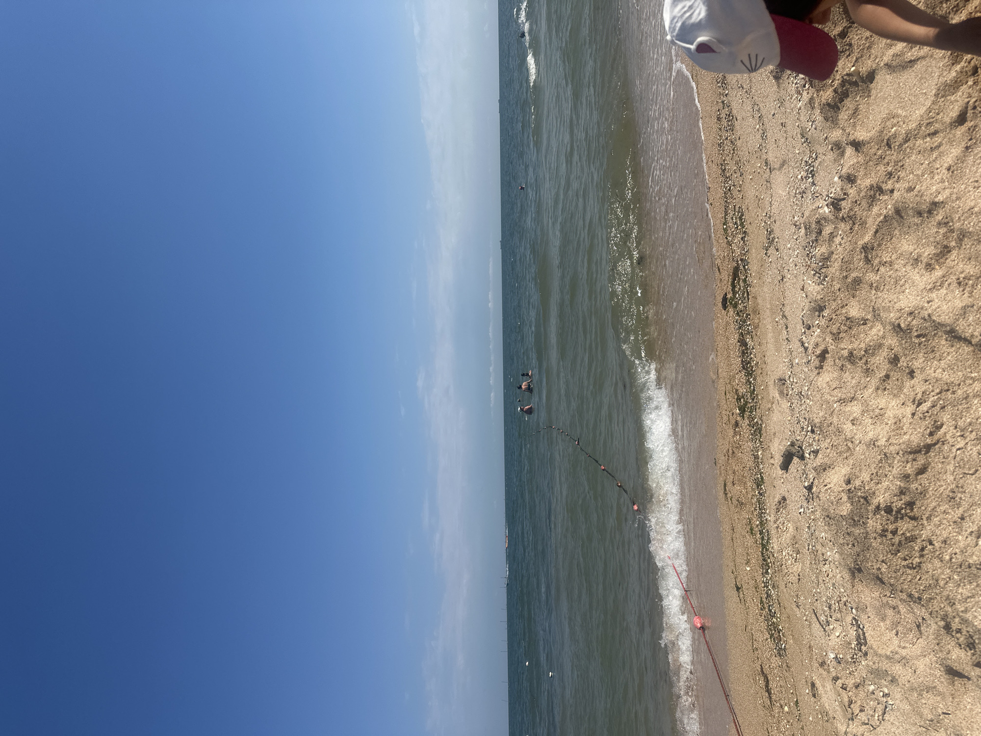 This is a medium shot of a beach in Golubitskaya, Russia, on a clear day. The sky is bright blue with a few scattered clouds. The sea is calm and a few people are visible swimming in the distance. A rope with buoys separates the swimming area from the rest of the sea. The sand on the beach is light brown and there are a few pieces of driftwood. In the foreground, a child wearing a white hat with cat ears and a pink brim is partially visible, looking down at the sand.