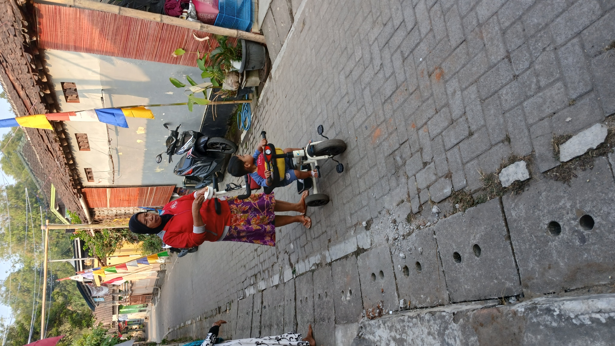 In a residential area of Mendut, Indonesia, a woman in a red jacket and patterned skirt stands beside a young child riding a tricycle. The street is paved with interlocking bricks, and a drainage ditch runs along the left side of the road. Several houses with terracotta tiled roofs and bamboo-woven walls line the street. Flags are displayed on poles in front of one of the houses. A motorcycle is parked in front of another house. The scene appears to be a typical day in the village, with people going about their daily activities.