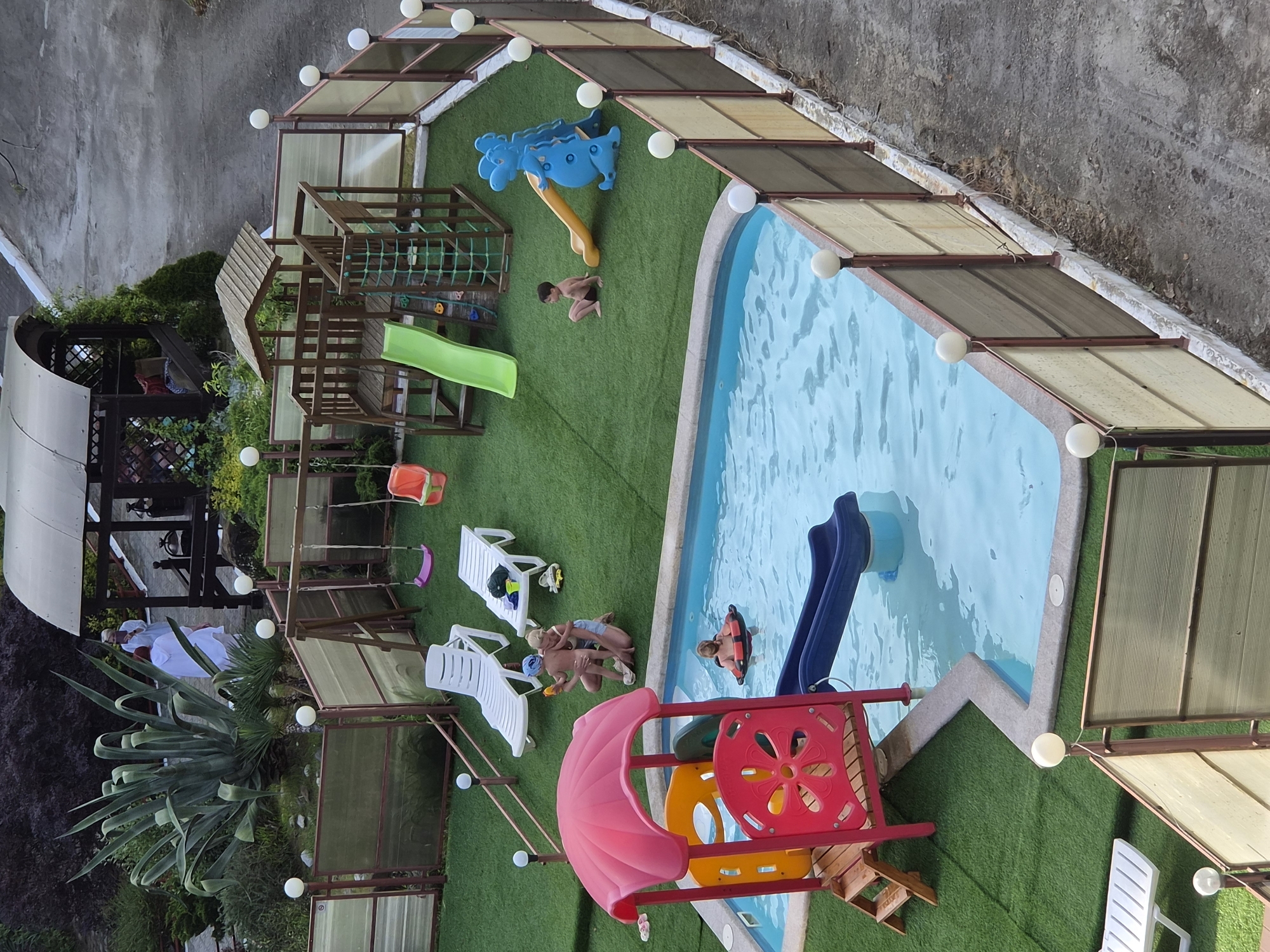 This image depicts a children's play area adjacent to a pool, likely in Sochi, Russia. The play area features artificial turf, a multi-level wooden play structure with a slide, a blue plastic climbing toy, a red and yellow playset with a pink umbrella, and a blue slide into the pool. Several people are present, including children and adults, some of whom are swimming in the pool, while others are lounging on white deck chairs. The area is enclosed by a fence with translucent panels and spherical lights. In the background, there's a covered seating area and a variety of plants.