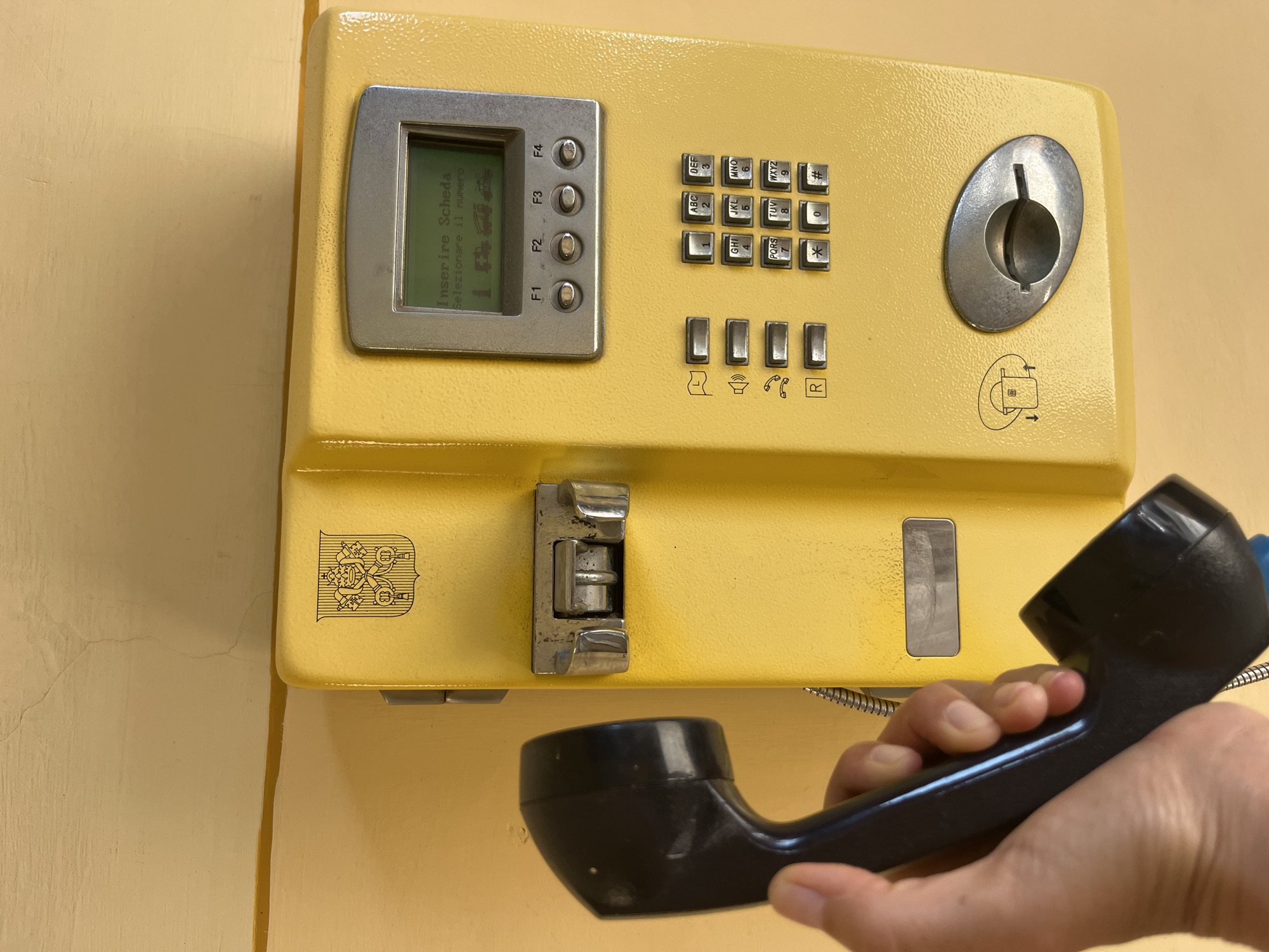 A yellow payphone is mounted on a light yellow wall. The payphone has a screen displaying "Inserire Scheda Selezionare il numero" and "15:00" and a keypad with numbers and symbols. The handset is being held in a person's hand, to the left of the payphone. The payphone has a coin slot and various function buttons. The wall has some texture and cracks. The payphone has an emblem at the top, possibly a coat of arms. The location is Città del Vaticano, Vatican City.