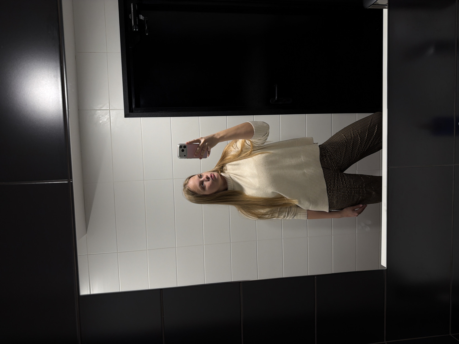 A young woman is depicted taking a mirror selfie in an indoor setting, likely in Izhevsk, Russia. She is positioned facing the mirror, holding a smartphone with a pink case in her left hand, which is aimed at her reflection. Her long, straight blonde hair flows over her shoulders. She is dressed in a light-colored, possibly cream or off-white, turtleneck sweater with ribbed cuffs and hem, paired with dark pants that feature a subtle, patterned design.
The background visible in the mirror reflection consists of white rectangular tiles on the wall. To the left of the woman's reflection, a large dark, potentially black, door or cabinet is seen. A bright, artificial light source creates a notable glare on the upper left section of the mirror. The mirror itself is framed by dark, possibly black, surfaces. The overall lighting suggests an indoor environment. The woman's expression is neutral as she looks towards the phone. No discernible text is visible on any objects or surfaces.