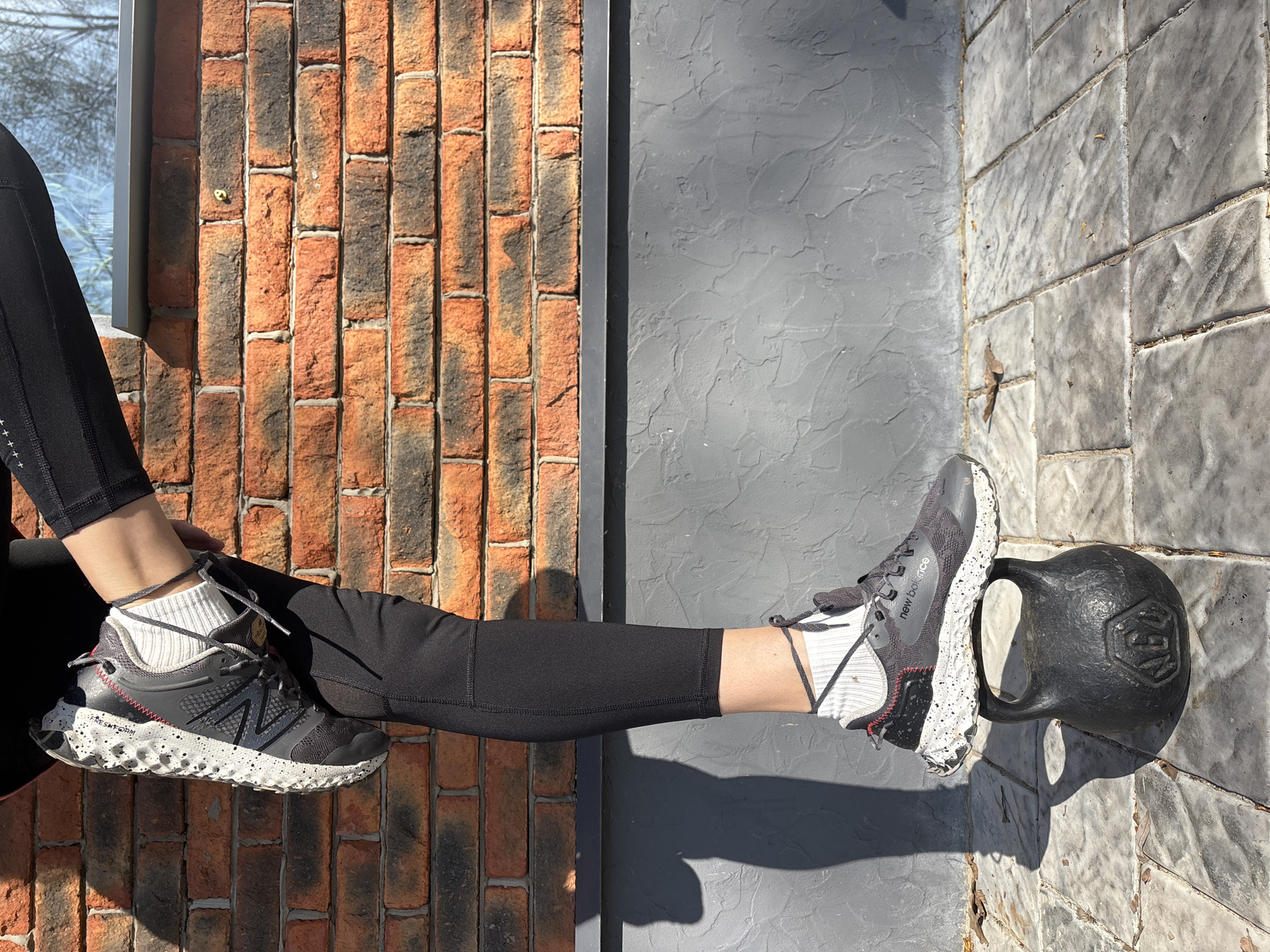 A person wearing black athletic leggings and New Balance athletic shoes sits with one leg resting on a kettlebell. The background features a brick wall, gray paneling, and a window with a reflection. The location appears to be outdoors, possibly in Chaikovsky, Russia.