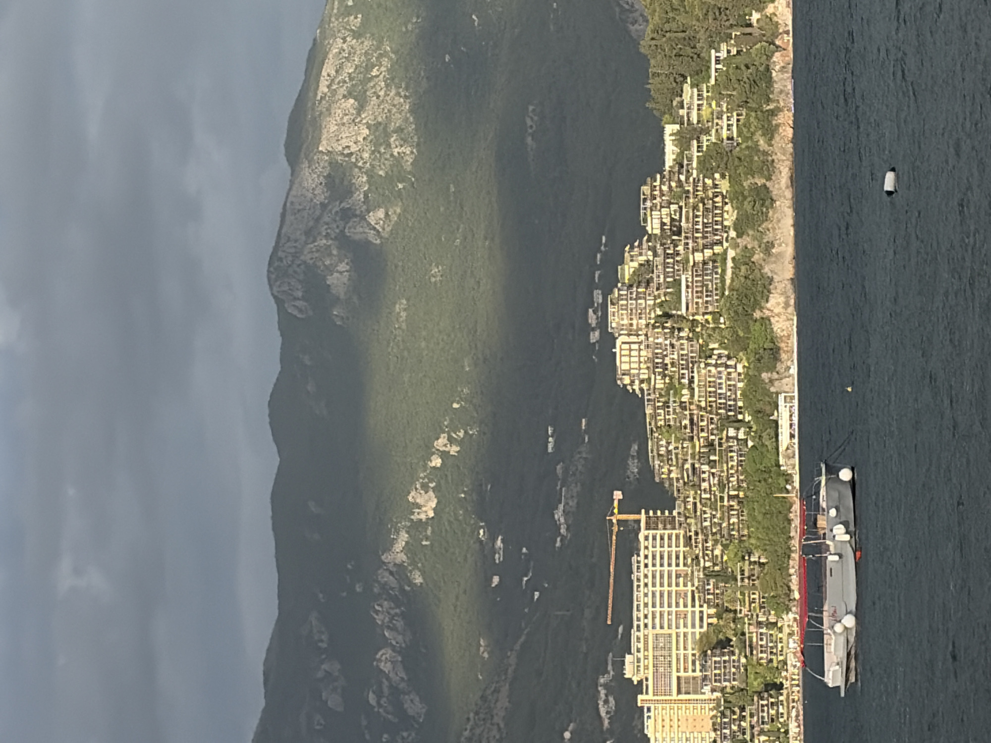 The image shows a coastal landscape in Budva, Montenegro. Dominating the view is a large mountain with varied terrain, partly covered in green vegetation. Along the coastline, there are numerous buildings clustered together, seemingly residential or hotel structures. A crane is visible among the buildings, suggesting ongoing construction. In the water, a large ship is docked near the coast. Further out, a single white buoy floats on the water. The sky is overcast with dark clouds on the left side of the image.