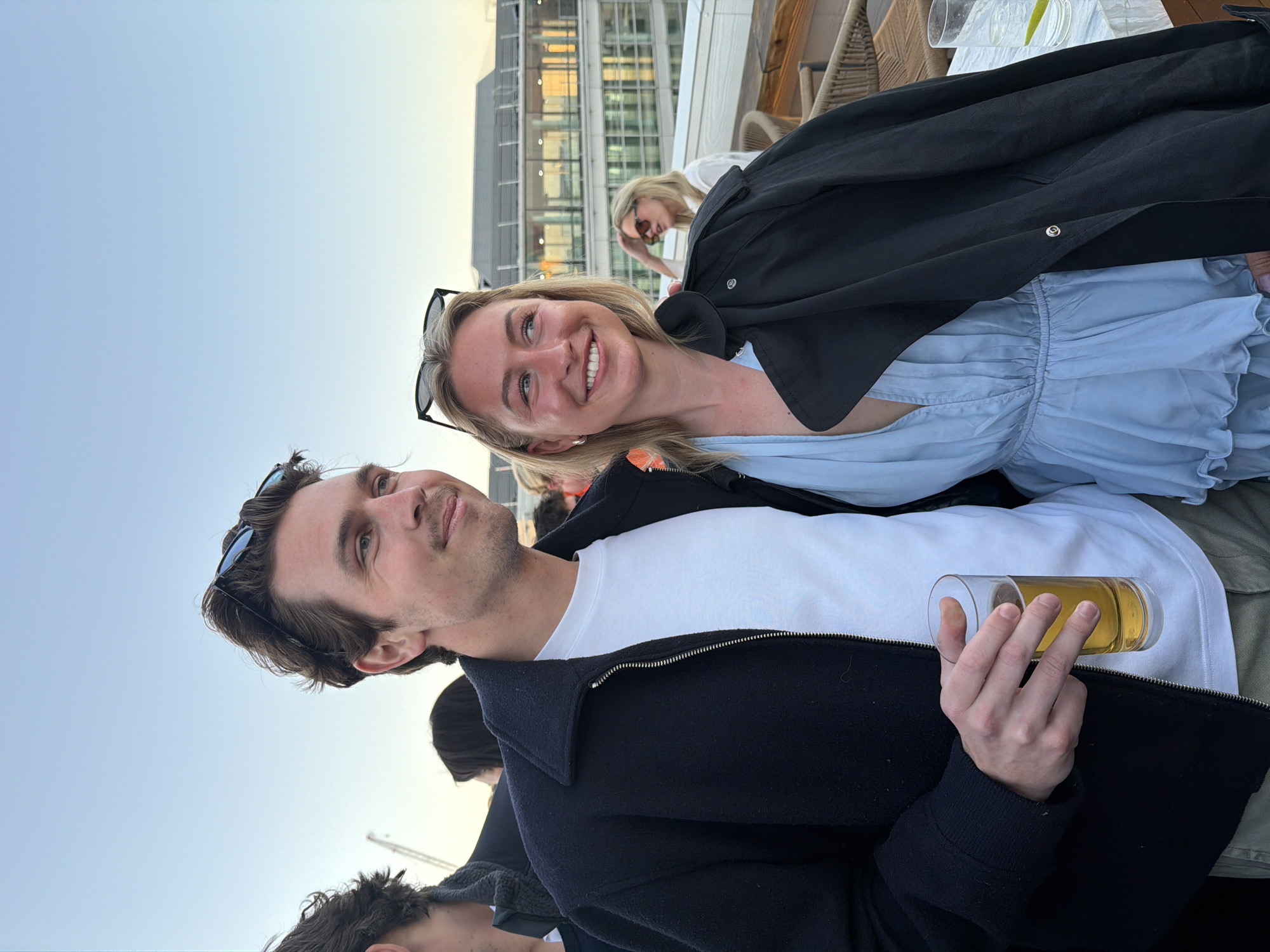 The image shows a man and a woman smiling for a photo outdoors, likely during the evening. The man is holding a glass of beer and wearing a white shirt and a dark jacket with sunglasses on his head. The woman is wearing a light blue top with a dark jacket draped over her shoulders and sunglasses on her head. They are standing on a rooftop or balcony, with a view of a modern building complex in the background. Other people are visible in the background, including another woman with sunglasses. Based on the architecture and the river view in the background, the location is likely in the city of London, United Kingdom.