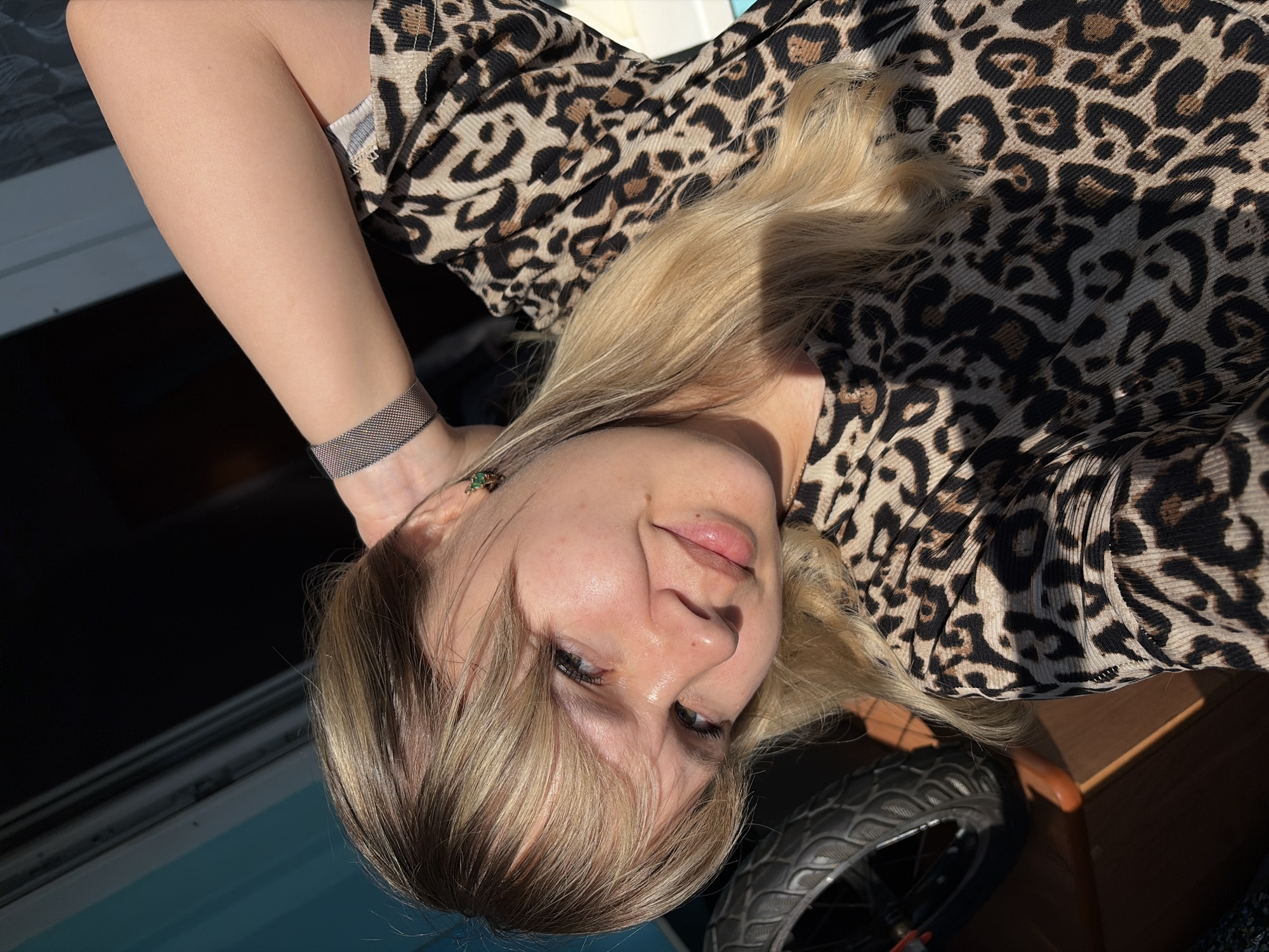 A young, light-skinned woman with blonde hair is pictured wearing a leopard-print garment and a mesh bracelet on her wrist. She appears to be seated near a window and a small wheel, potentially part of a chair or stroller, is visible in the lower right corner. The location of the image is not identifiable as Chaikovsky, Russia, based solely on visual elements.