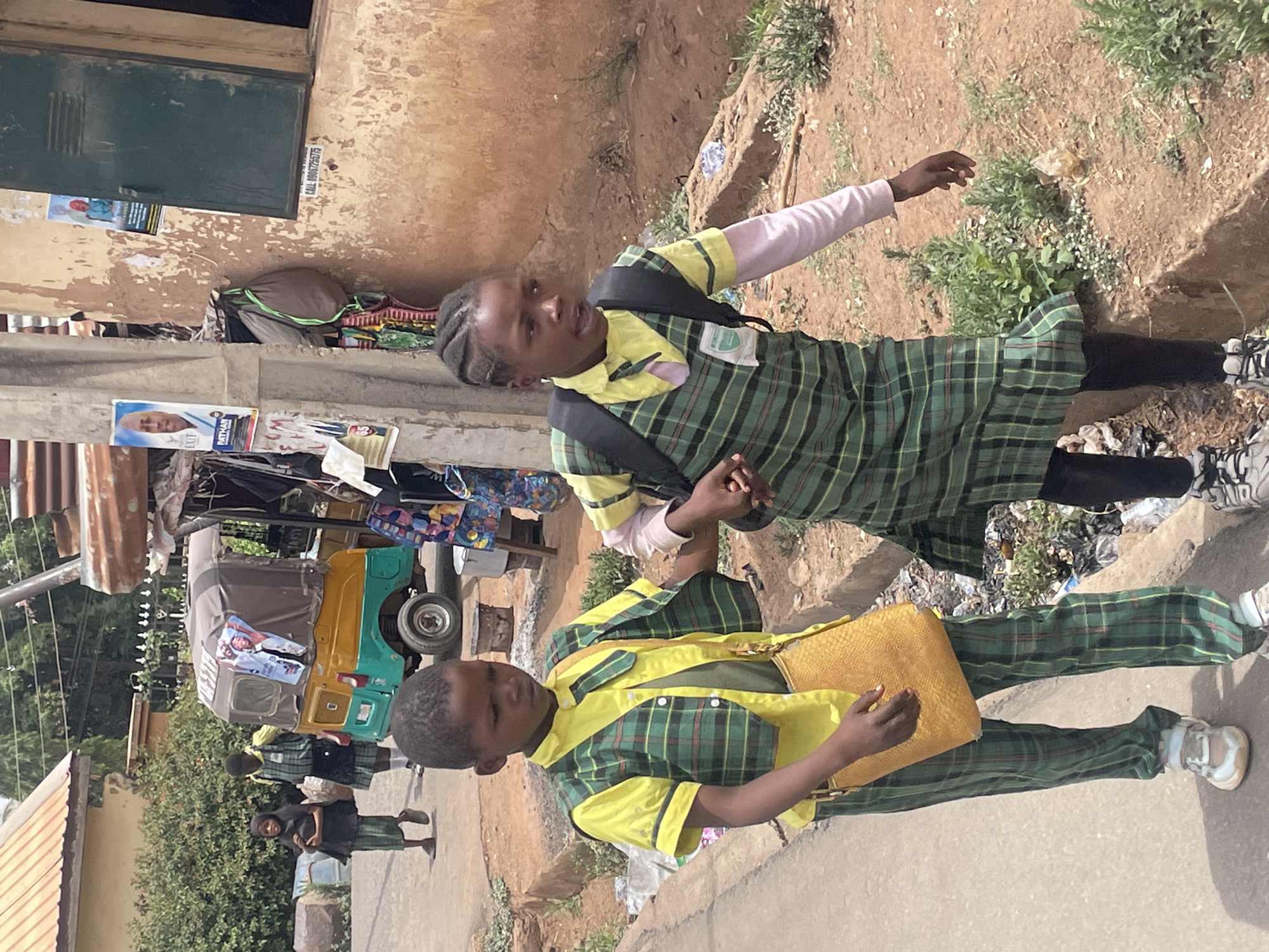 This image captures an outdoor scene in Jos, Nigeria, featuring two school-aged children walking hand-in-hand along a path. The child on the left wears a green and yellow plaid dress uniform with a black backpack and light pink long sleeves visible underneath, and looks towards the viewer. The child on the right wears matching plaid pants and shirt, holding a yellow textured handbag, and looks slightly ahead.
The setting is an outdoor street or pathway, characterized by a mix of dirt and paved surfaces, with scattered debris and sparse vegetation. Buildings line the left side, one featuring a dark green metal shutter and a wall with peeling, faded yellow paint. A visible sticker on this wall reads "CALL 08067256775", confirming a Nigerian context. Further down the path, a yellow and green three-wheeled auto-rickshaw (commonly known as a keke Napep in Nigeria) has posters affixed to its side, showing a man in a white cap and visible text including "PATHWAY" and "EXIT". Other people, including another child in a similar uniform and a black head covering, are visible in the background.
The scene is bathed in bright daylight, suggesting midday or early afternoon under clear weather conditions. The children's expressions are neutral, portraying a typical daily commute, likely to or from school, in this local neighborhood in Jos, Nigeria.
