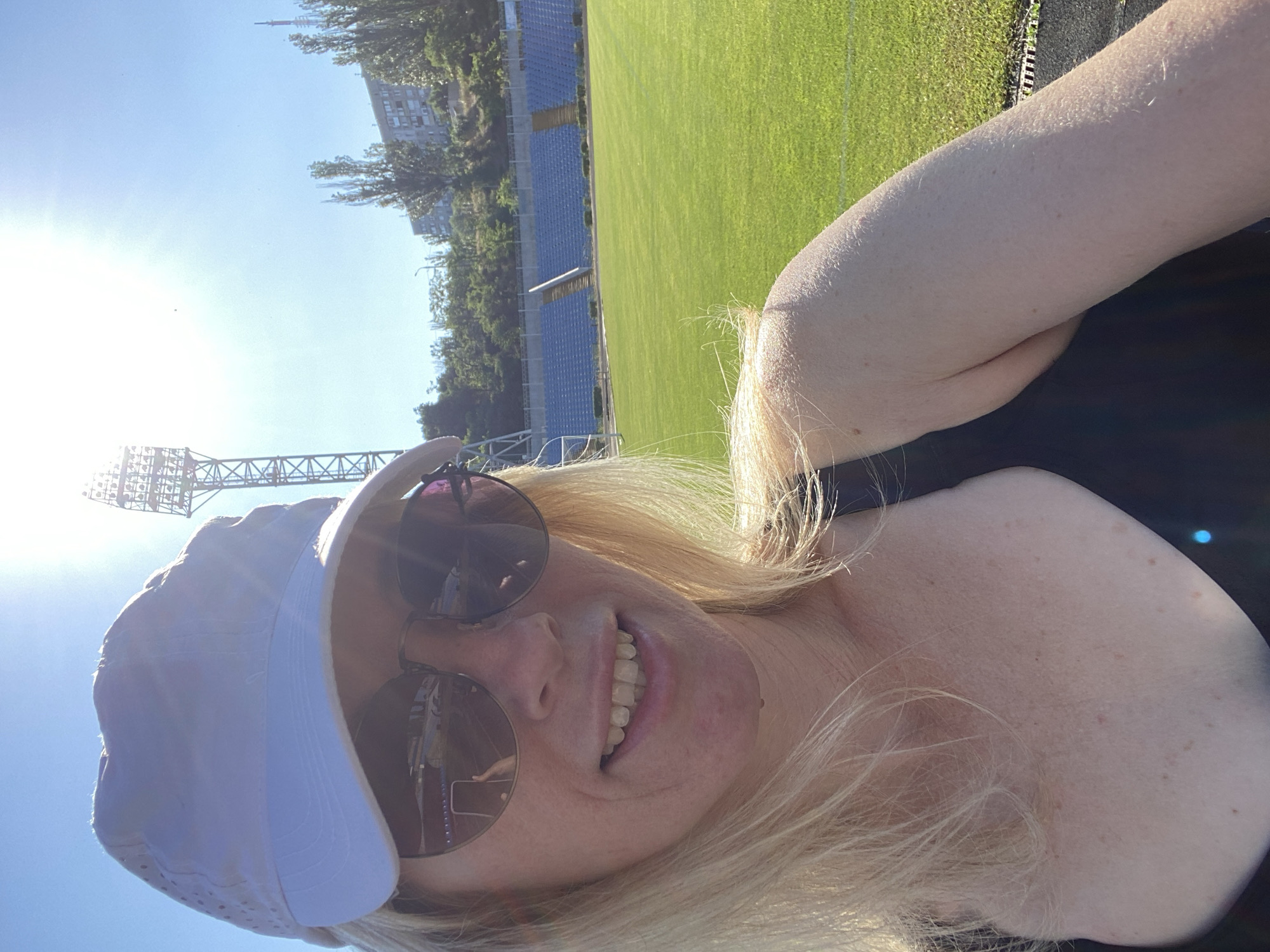 A selfie taken from a slightly low angle shows a blonde woman with freckles on her skin, wearing a white baseball cap and sunglasses. She has a slight smile. The background suggests a sunny day at a sports stadium, with a green field, blue seating, and a tall light tower. Some trees and buildings are visible in the distance. The woman's arm and shoulder are visible in the foreground, with the sun glinting off her skin.