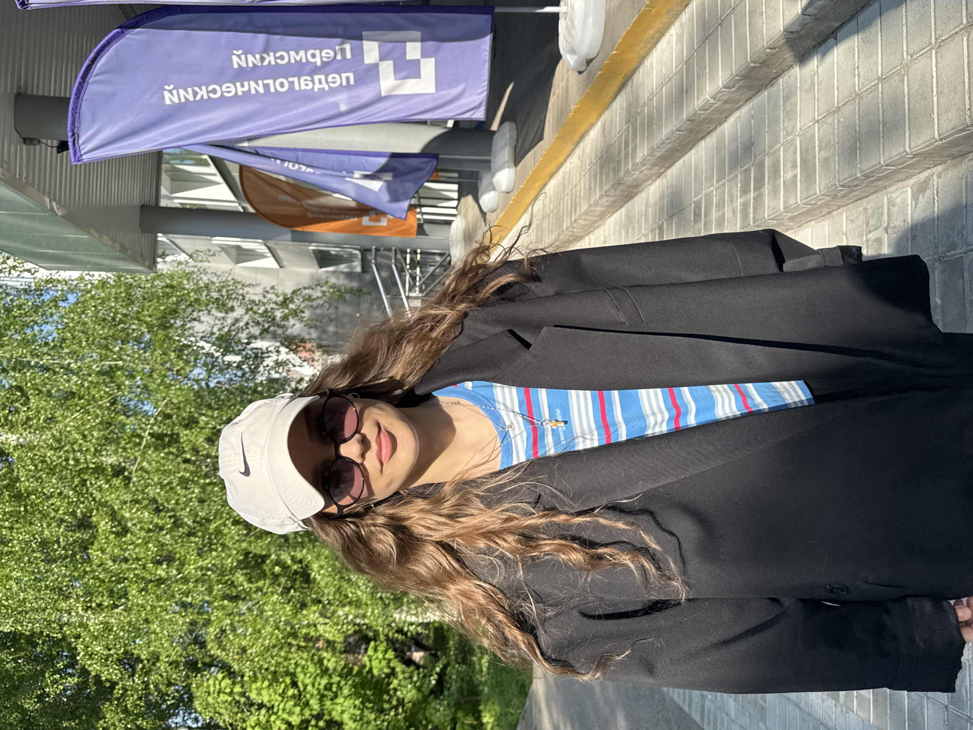 The image shows a woman wearing a white Nike baseball cap, sunglasses, a blue and white striped shirt, and a black blazer. She is standing outdoors. Trees are visible in the background. The backdrop includes banners written in Russian. Based on the banners, the location is likely in Perm, Russia. The specific location cannot be determined from the image alone.