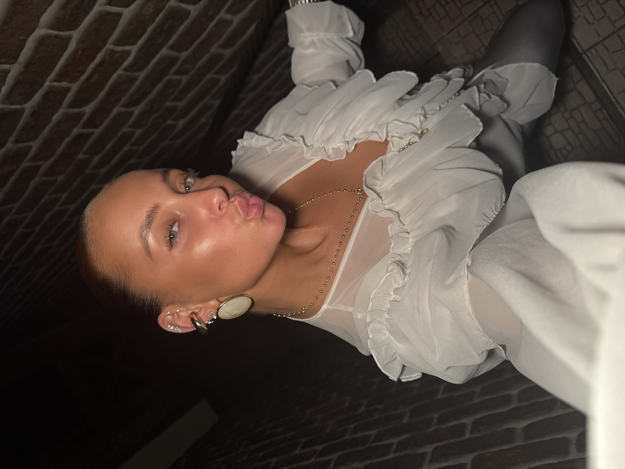 The image shows a young woman posing for a selfie. She is wearing a white ruffled blouse and gray leggings. She has her hair pulled back and is wearing large gold earrings and a delicate gold necklace. Her facial expression is a duck face, and she is looking into the camera. The background appears to be a brick wall, suggesting an indoor setting, possibly a room or hallway. The lighting is dim, creating shadows. The image does not provide any specific details that would allow for identification of a location in Chaikovsky, Russia.