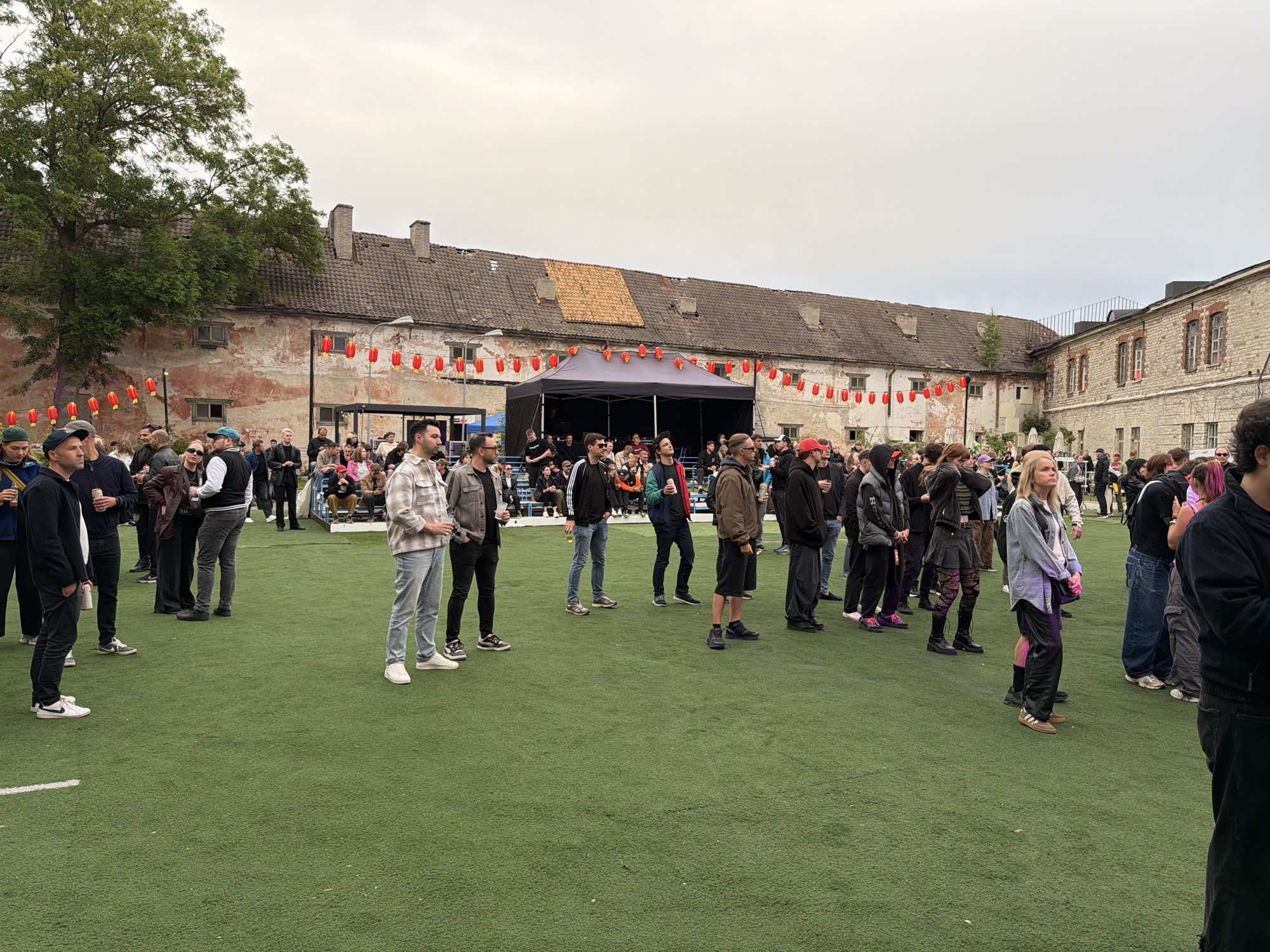 The image depicts an outdoor gathering in Tallinn, Estonia, likely at the Linnahall area, a former limestone production facility and a concert venue. A large crowd of people, predominantly young adults, are standing on a bright green artificial turf field, facing a black tent set up as a stage. Red paper lanterns are strung along the buildings in the background, which appear to be old industrial structures with weathered facades and tiled roofs.
The people in the foreground are casually dressed in everyday attire, including jeans, t-shirts, jackets, and sneakers. Many are looking towards the stage, suggesting they are attending an event, possibly a concert or festival. Some individuals are holding drinks or standing in small groups. The overall atmosphere seems relaxed and informal. The sky is overcast, indicating cloudy weather.
