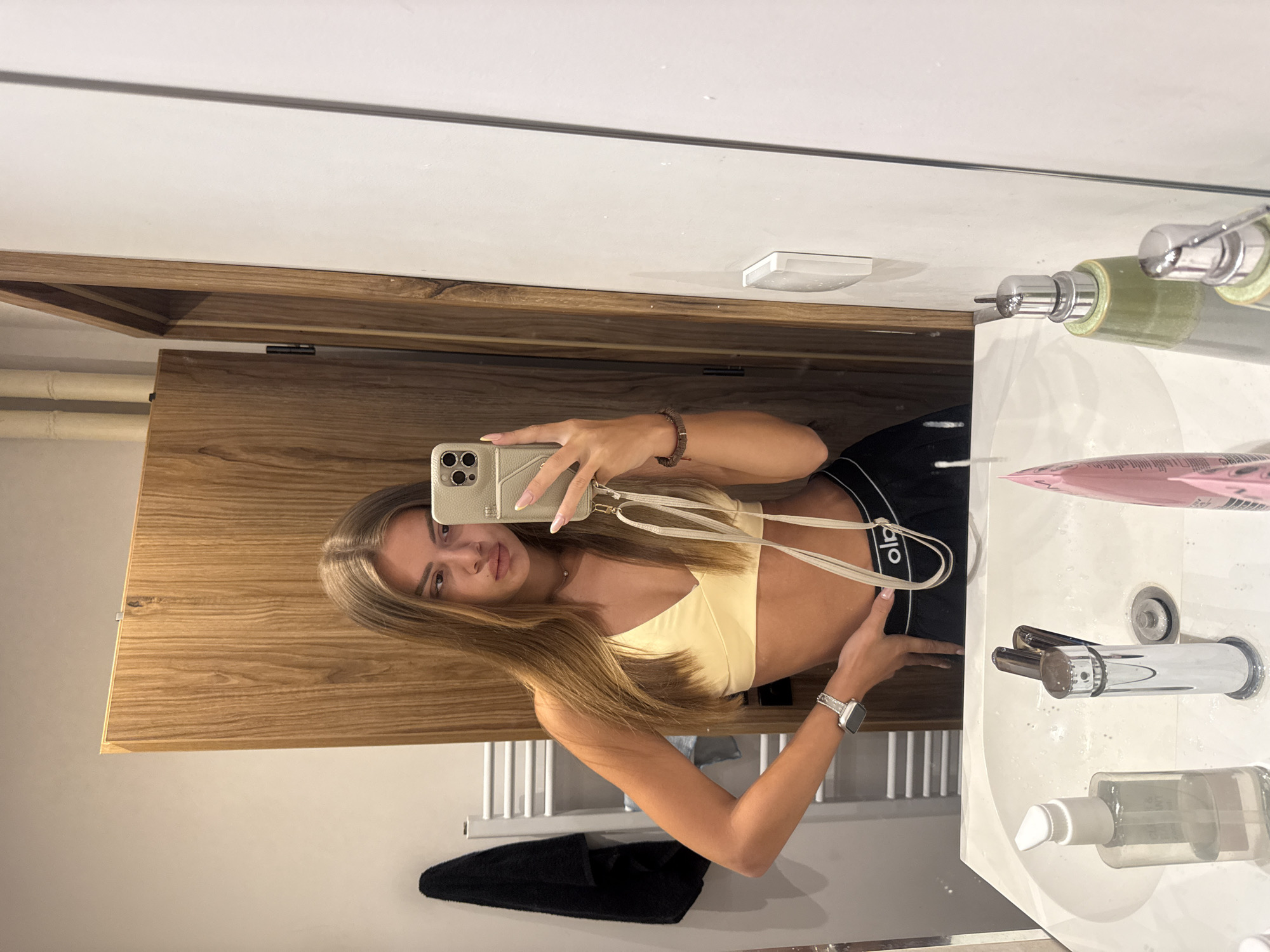 A young woman with long, brown hair takes a mirror selfie in a bathroom. She is wearing a light yellow sports bra and black athletic shorts with a white waistband. Her left arm is bent, resting on her hip, and her right hand holds a phone to take the picture. A silver smartwatch is visible on her left wrist. To her left, a towel rack with a black towel hangs on the wall. In front of her, a white sink with chrome faucets and various toiletries is visible. The bathroom appears to be modern, with wooden accents and white tiled walls. The scene is likely in a residential bathroom within the city of Plovdiv, Bulgaria, given the context provided.