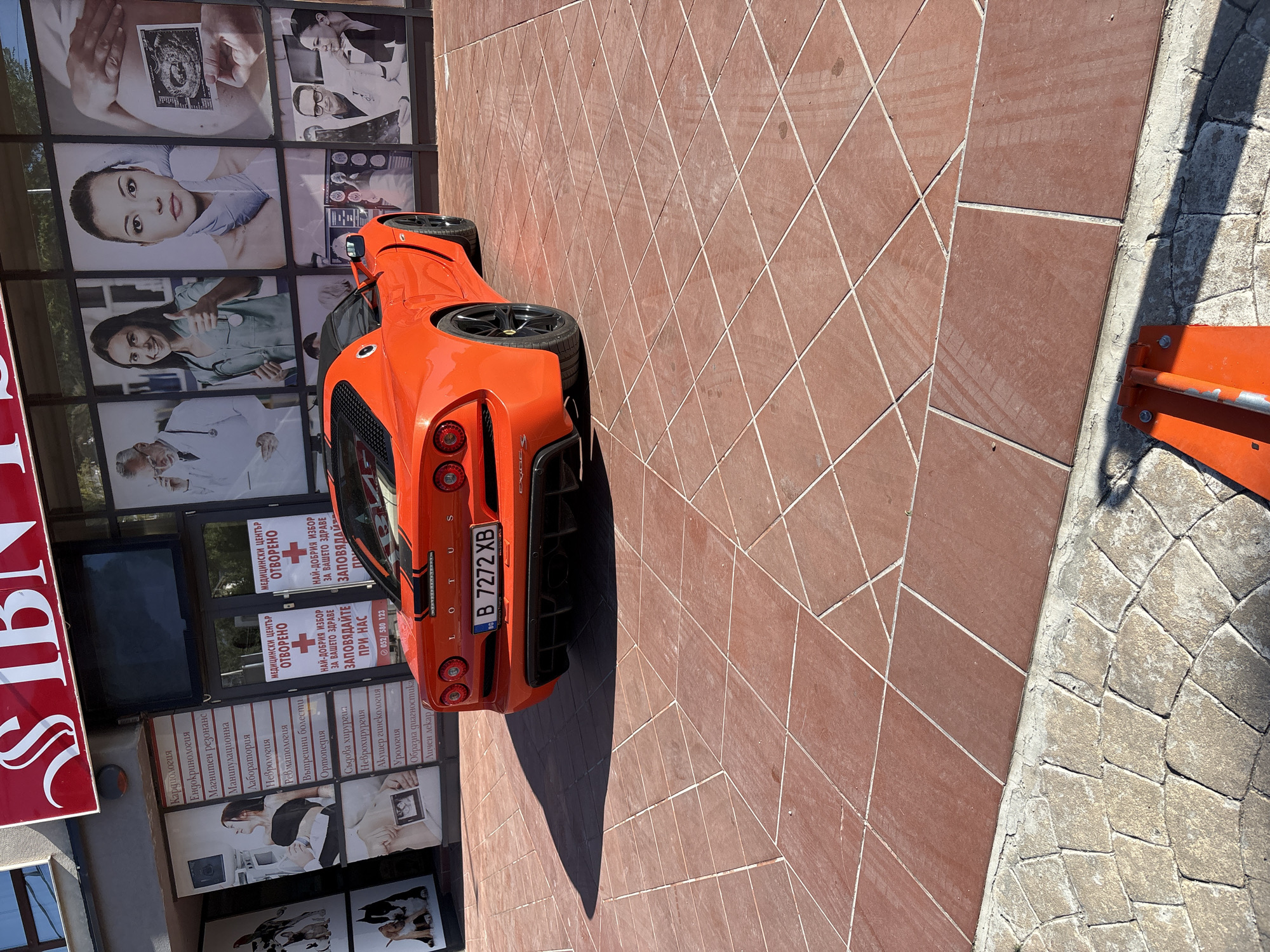This image features an orange Lotus sports car parked in front of a building with several glass panels displaying medical information and advertisements. The building appears to be a medical center, as indicated by the text visible on the glass, which includes names of medical specialties like Cardiology, Neurology, and Urology, as well as phrases like "Medical Center Open" and "Your Health." The car's license plate is visible, reading "B 7272 XB," suggesting it is registered in Bulgaria.
The scene is outdoors, likely on a sunny day, given the bright lighting and the sharp shadow cast by the car onto the tiled ground. The tiles are reddish-brown and arranged in a diamond pattern. A cobblestone path is visible to the right of the tiled area, bordering a concrete edge. The overall impression is a vibrant, modern setting, with the sleek sports car contrasting with the more utilitarian facade of the medical building. No people are clearly visible in the frame, but some of the advertisements on the building show images of people, likely medical professionals or patients. The time of day appears to be daytime, and the weather seems fair.