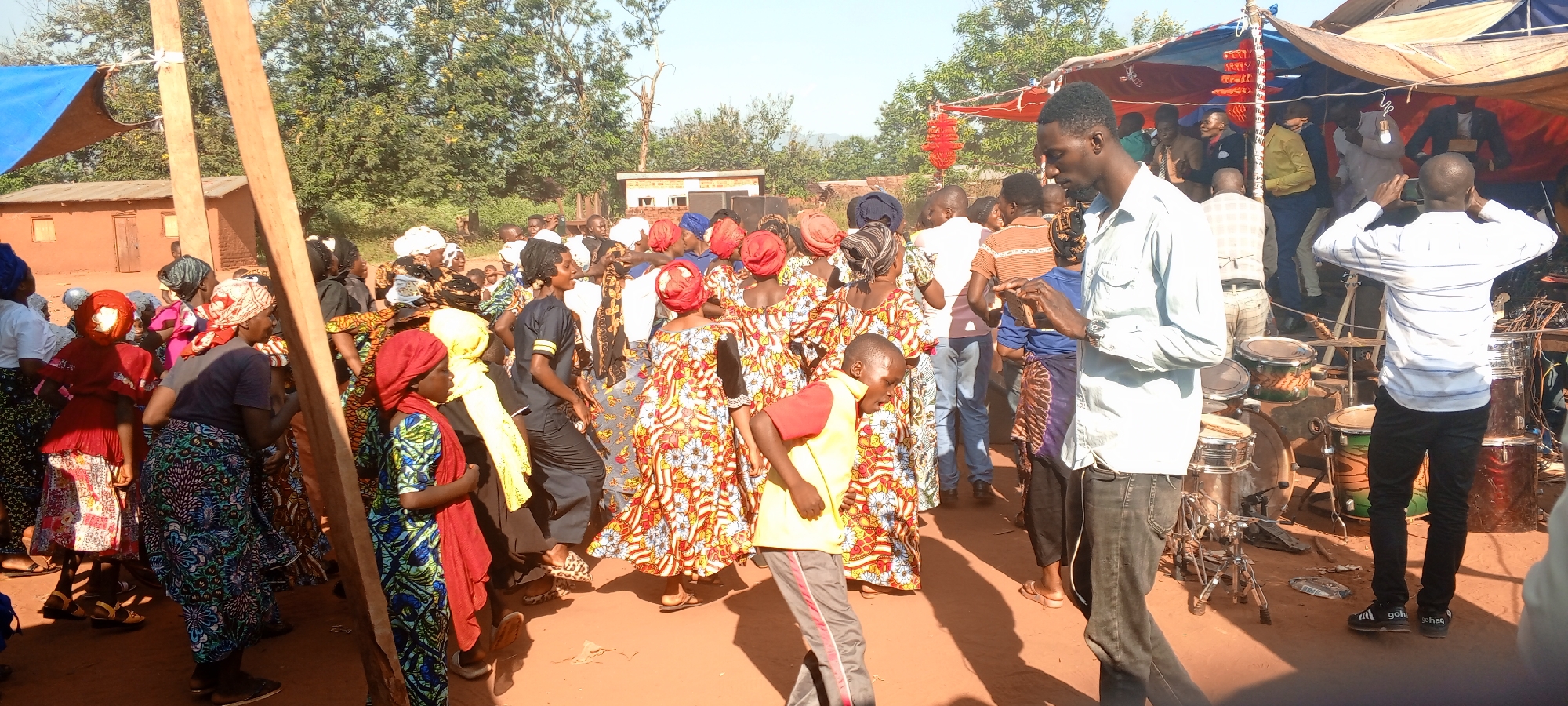 Captured in Ngalula, Democratic Republic of the Congo, the image reveals a lively outdoor gathering. A group of individuals, predominantly women adorned in vibrant, patterned dresses and headwraps, occupy the center, seemingly engaged in dancing or celebration. To the right, a man in a light blue shirt is focused on a phone screen, while another man wearing a striped white shirt and black pants stands by a drum set, possibly involved in recording or observing the event. In the background, a tent-like structure provides shade, and various buildings and trees indicate the setting. The foreground is characterized by the reddish-brown ground, typical of the region.