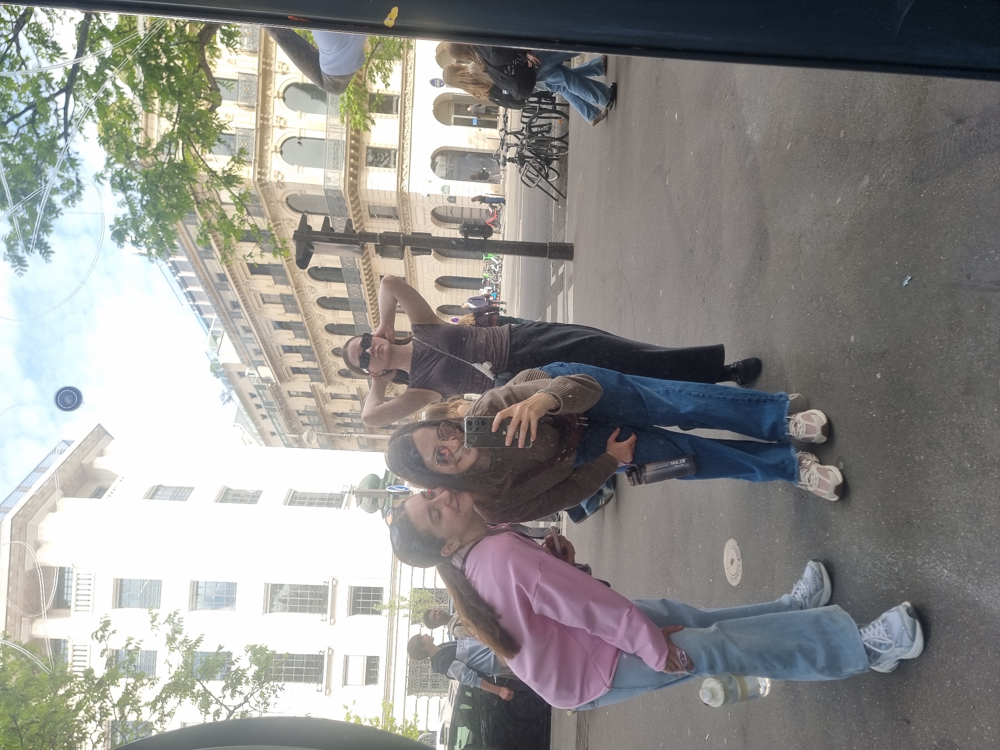 The image captures a vibrant street scene in Paris, France, reflected on a shiny surface, likely a window or glass panel, giving it a somewhat distorted and dreamy quality. The perspective is vertical, but the reflected scene shows a horizontal street.
Three young women are prominently featured in the reflection, appearing to be posing or interacting. In the foreground, a woman with long, braided dark hair, wearing a pink long-sleeved top and light blue jeans, leans slightly toward another woman. This second woman, wearing a brown sweater, blue jeans, and sunglasses, holds a smartphone up, seemingly taking a selfie or group photo. The text "FLASH" is visible in reverse on her phone case, and "ELAH" (likely a mirrored brand name) is on a dark bottle she carries. Behind them, a third woman, dressed in a dark brown top and black pants, also wearing sunglasses, stands with one hand behind her head in a relaxed pose. All three appear to be in good spirits, enjoying a sunny day.
The background shows classic Parisian architecture with multi-story buildings featuring ornate facades, numerous windows, and some balconies. Green trees and a bright, partly cloudy sky are reflected above the buildings. The street below the reflection shows a sidewalk with parked bicycles and other pedestrians further in the distance. The ground is paved, and a circular metal utility cover is visible. The bright lighting indicates it is daytime, likely a clear and sunny day.