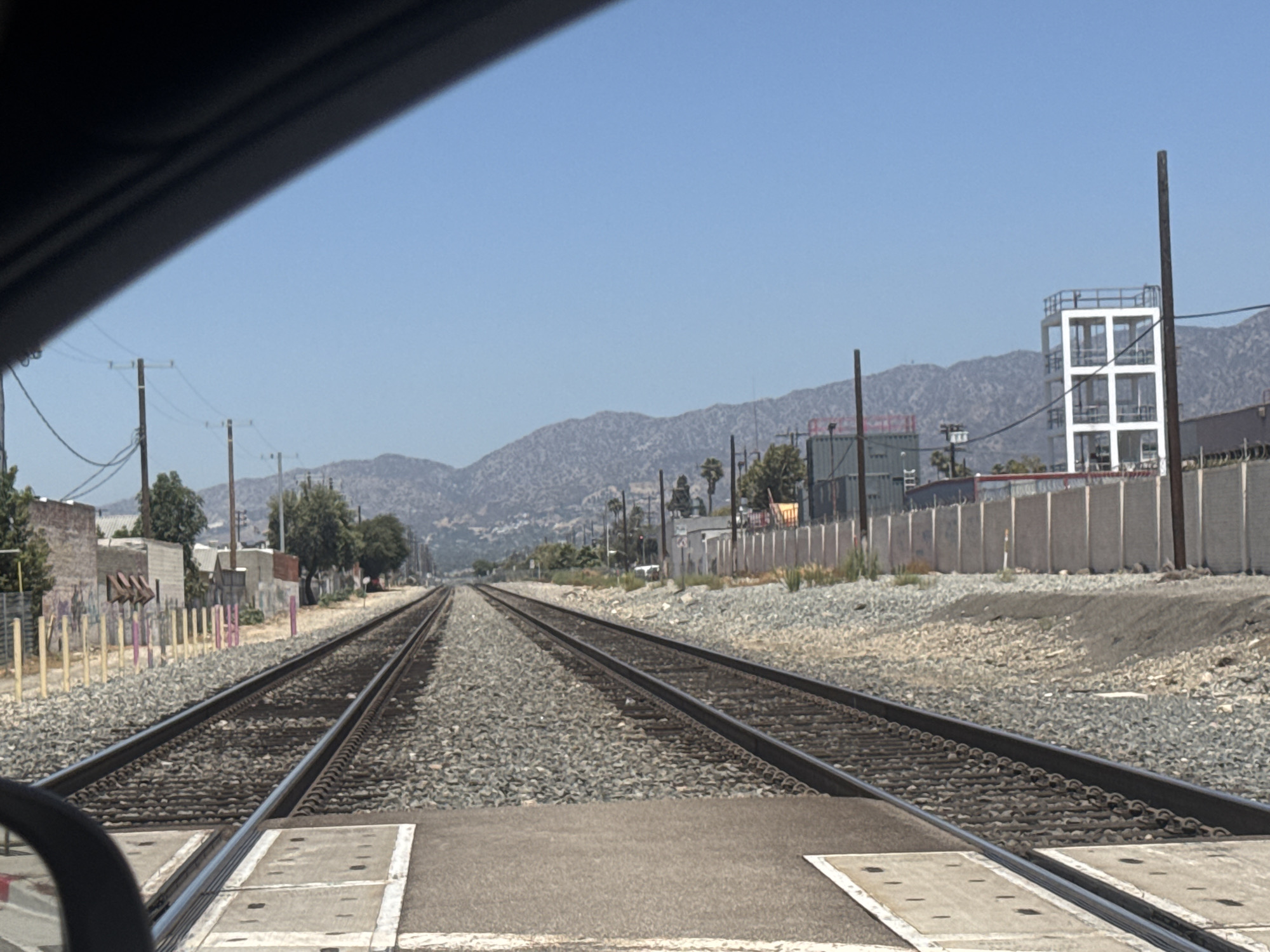 A daytime, eye-level, medium shot captures a set of railroad tracks in an industrial area. The tracks stretch into the distance, flanked by ballast and a gravel-covered shoulder. On the left, low-rise buildings with some graffiti are visible, along with a fence. On the right, a concrete wall runs parallel to the tracks, and beyond it, shipping containers and a multi-story white structure are present. In the background, a range of mountains under a clear blue sky completes the scene. A car's rearview mirror is partially visible in the lower left corner.