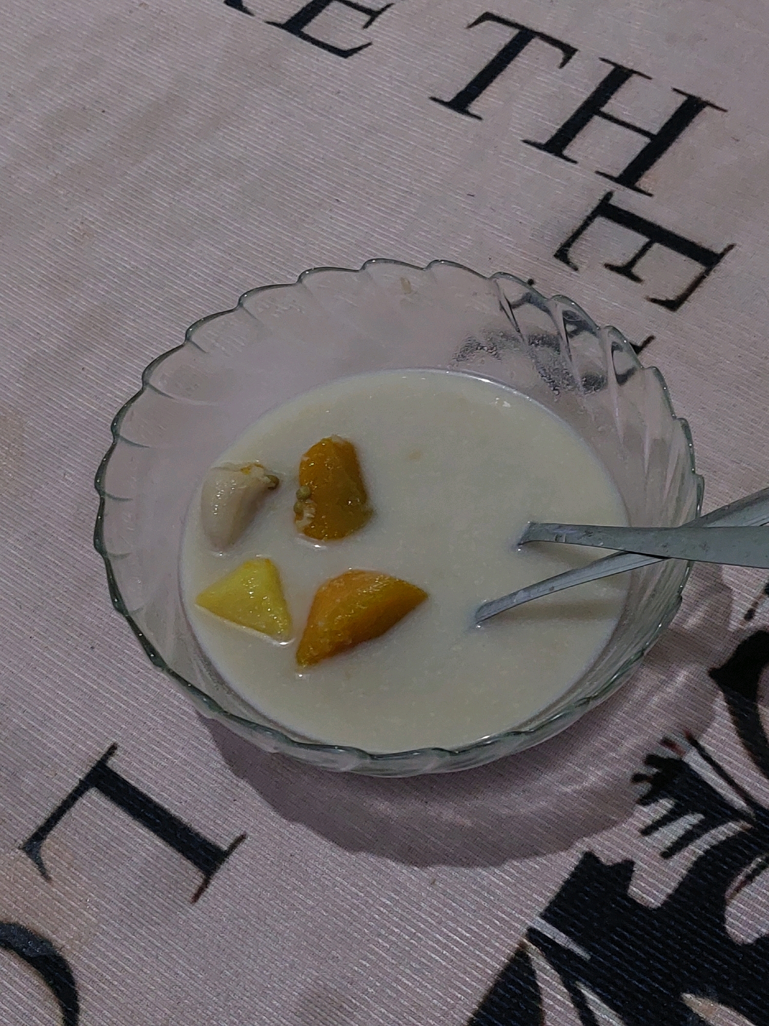 A clear, fluted glass bowl filled with a white liquid and several pieces of fruit is positioned on a pinkish-white patterned tablecloth. The fruit pieces are of varying shapes and colors, appearing to be chunks of yellow and orange fruit, with one light-colored, elongated piece. Two metal spoons are resting in the bowl, one partially submerged and the other crossed over it. The tablecloth has a repeating pattern of large, black, serif letters, with parts of the letters "T", "H", and "E" visible in the image, suggesting text. The overall lighting suggests an indoor setting, possibly a dining area. The image is an overhead shot, capturing the contents of the bowl and the immediate surrounding tablecloth. No specific location in Sarangan, Indonesia is identifiable from the visual information presented.