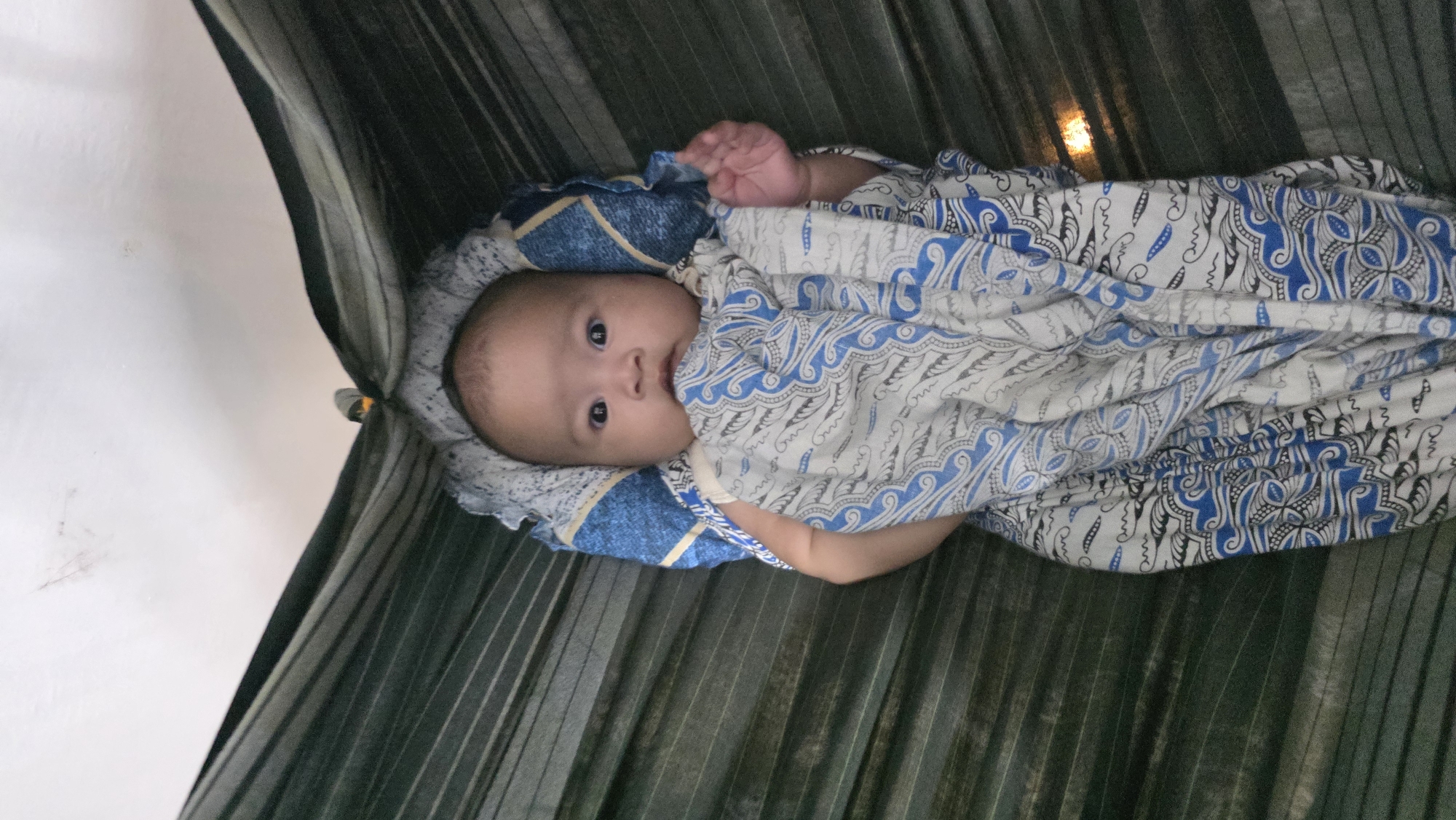 A baby is lying in a cot or bassinet. The baby is wrapped in a white blanket with a blue swirling pattern. The baby's face is visible, and it is looking directly at the camera. The baby's left hand is raised. The cot is lined with a blue and white patterned fabric, and the background appears to be made of dark green corrugated plastic or metal. This scene could be in Lhokseumawe, Indonesia, based on the user's query, though the image does not explicitly show any identifiable location information.