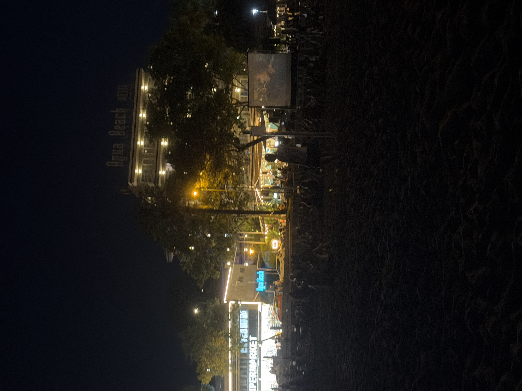 In the city of Calvià, Spain, a nighttime scene unfolds on a sandy beach where an outdoor cinema event is taking place. A large projection screen displays a movie to an audience seated on the sand. In the background, a brightly lit supermarket with the sign "IPERMARKET" is visible, along with other businesses and buildings, including the "Aqua Beach HOTEL" with illuminated signage. Trees and streetlights dot the landscape, casting a warm glow on the scene. The beach is covered in sand, with some areas appearing slightly rippled. The overall atmosphere is relaxed, with people gathered to enjoy a movie under the stars.