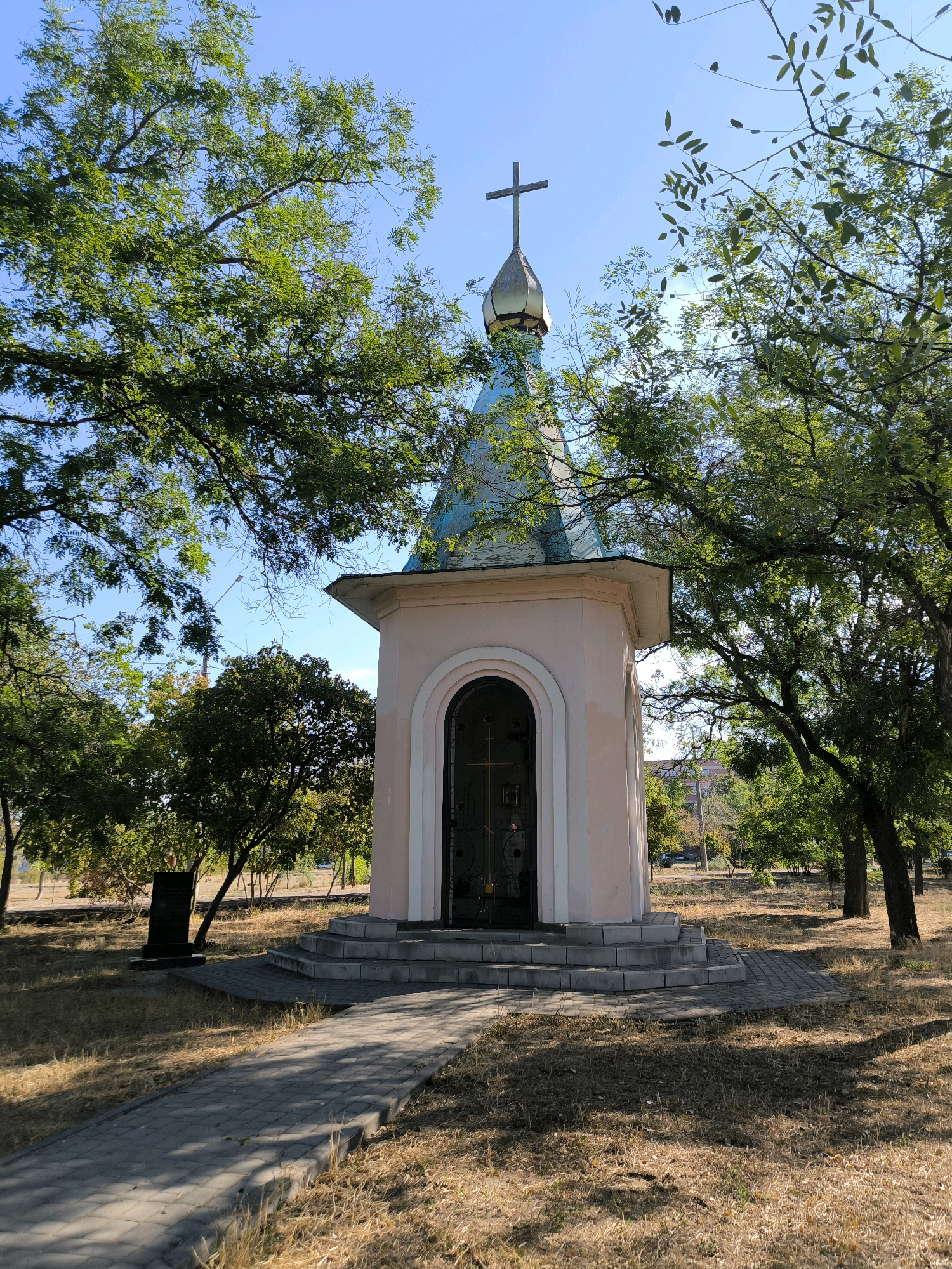The image shows a small, light pink chapel with a blue-green domed roof and a cross on top. It is situated in a park-like setting with dry grass and mature trees surrounding it. A paved walkway leads up to a set of stone steps in front of the chapel's entrance, which is a dark metal door. To the left of the chapel, a dark rectangular monument stands upright in the grass. The sky is clear and blue. The scene appears to be in Mykolaiv, Ukraine, likely a place of religious significance or a memorial.