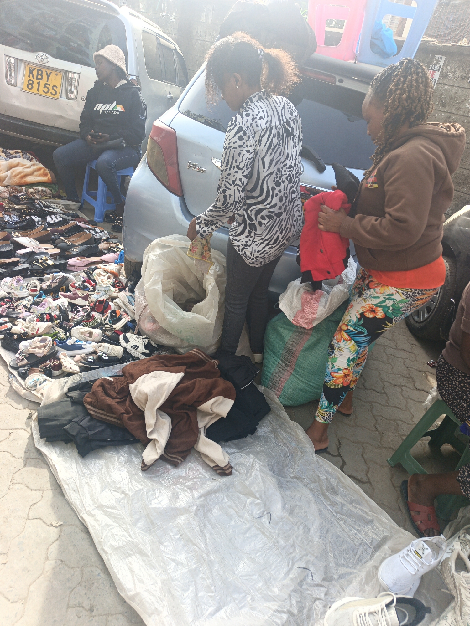 The scene shows a street market in Nairobi, Kenya. Several people are present, presumably buying and selling goods. One woman, wearing a zebra-patterned shirt and jeans, appears to be sorting through items. Another woman, dressed in a brown hoodie and floral leggings, is holding a red jacket. There's also a woman wearing a black hoodie and a hat seated on a chair near a vehicle with the license plate KBY 815S.
The market setup includes a large number of shoes spread out on the ground alongside a white plastic tarp, bags filled with clothing, and piles of clothes. The environment suggests a bustling, possibly informal, marketplace. Several vehicles are parked nearby, indicating the urban setting.