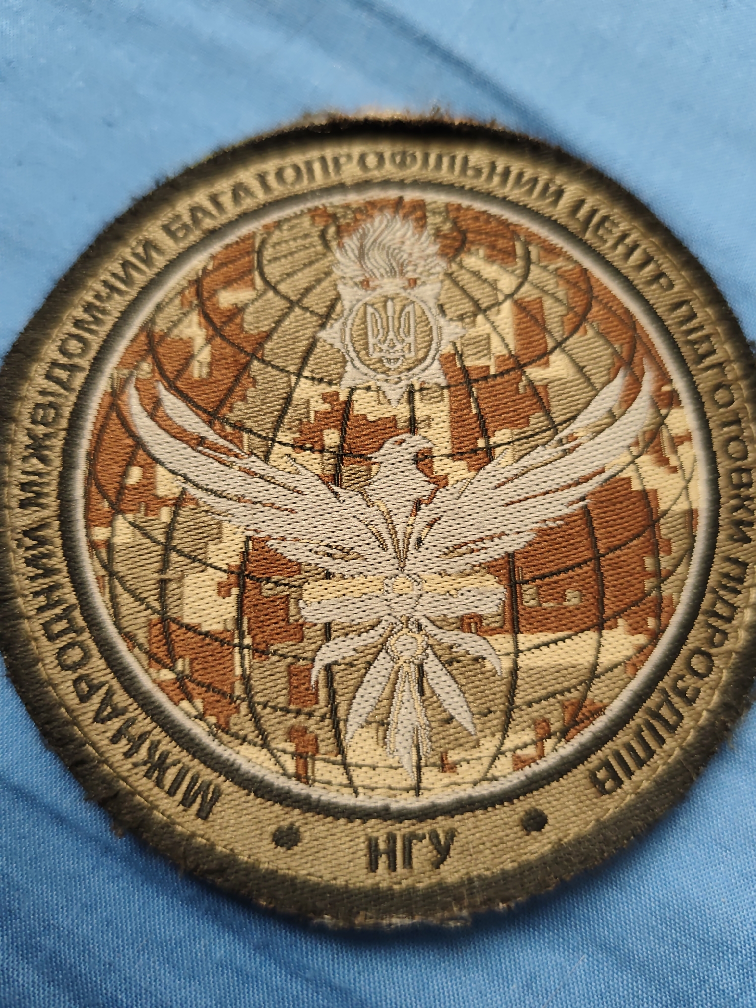 The image shows a circular patch with intricate embroidery. The patch features a globe with a camouflage pattern, a stylized bird with outstretched wings, and the Ukrainian trident emblem at the top. The outer rim of the patch contains text in Cyrillic script. Below the central design, the letters "НГУ" are embroidered. The patch is presented against a blue fabric background. Based on the presence of "НГУ," which stands for the National Guard of Ukraine, and the imagery commonly associated with Ukrainian military or service insignia, this patch likely belongs to a member or organization within the National Guard of Ukraine. Given the request for specific location details in Stare, Ukraine, and the absence of any visual information within the image that directly indicates this location, no definitive statement about its presence in Stare can be made from the image itself. The image solely captures the patch and its immediate background.