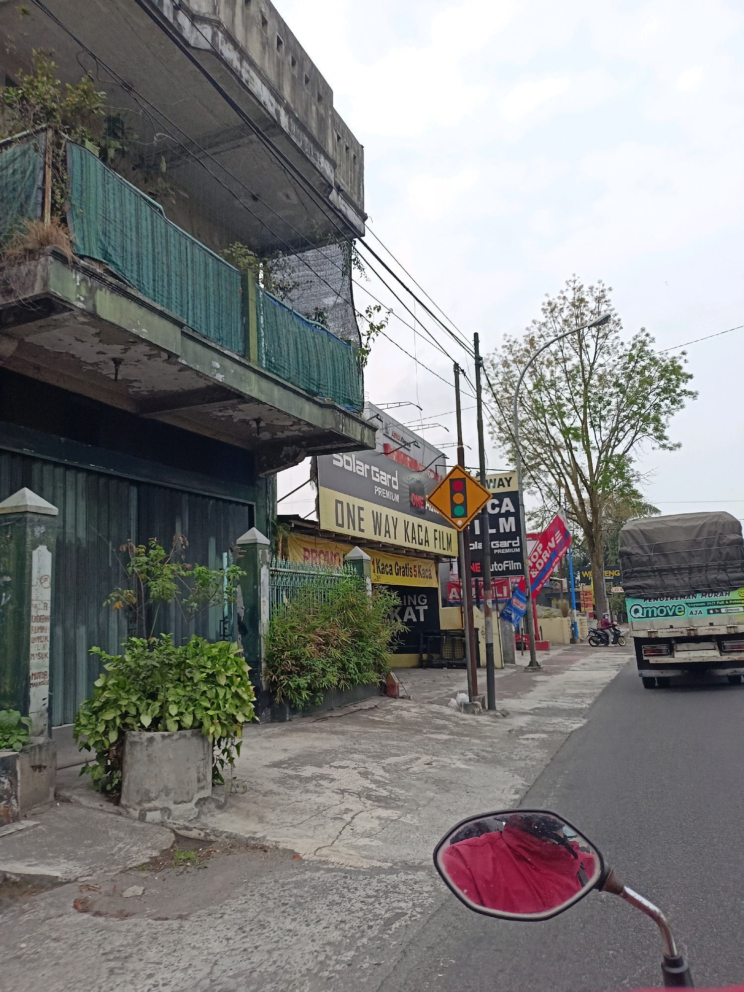 The image depicts a street scene in Magelang, Indonesia, on an overcast day. In the foreground, a motorcycle's side mirror reflects a person wearing a red jacket, suggesting the photo was taken from a motorcycle in motion. To the left, an aged, multi-story building with green tarps covering its balconies is visible. Adjacent to the building, a rolling metal gate stands closed. A concrete planter with a lush green plant sits on the sidewalk.
The sidewalk leads to a row of businesses, indicated by various signs. Prominently displayed are signs for "SOLAR GARD PREMIUM" and "ONE WAY KACA FILM," suggesting a shop selling car window film and related services. Other signs include "PROMO," "KACA GRATIS 5 KACA," and "TOKO AND DRIVE." A traffic signal with red, yellow, and green lights is positioned on a pole near the businesses.
Further down the street, trees line the sidewalk, with one particularly large tree with sparse foliage. Several utility poles and wires crisscross overhead. A truck with a covered cargo bed is moving away from the camera on the right side of the road. The truck has "PENGIRIMAN MURAH" (cheap shipping) and "Qmove" branding. A lone motorcyclist is also visible on the road, heading in the same direction as the truck. The overall environment appears to be a commercial area with a mix of old and newer structures.