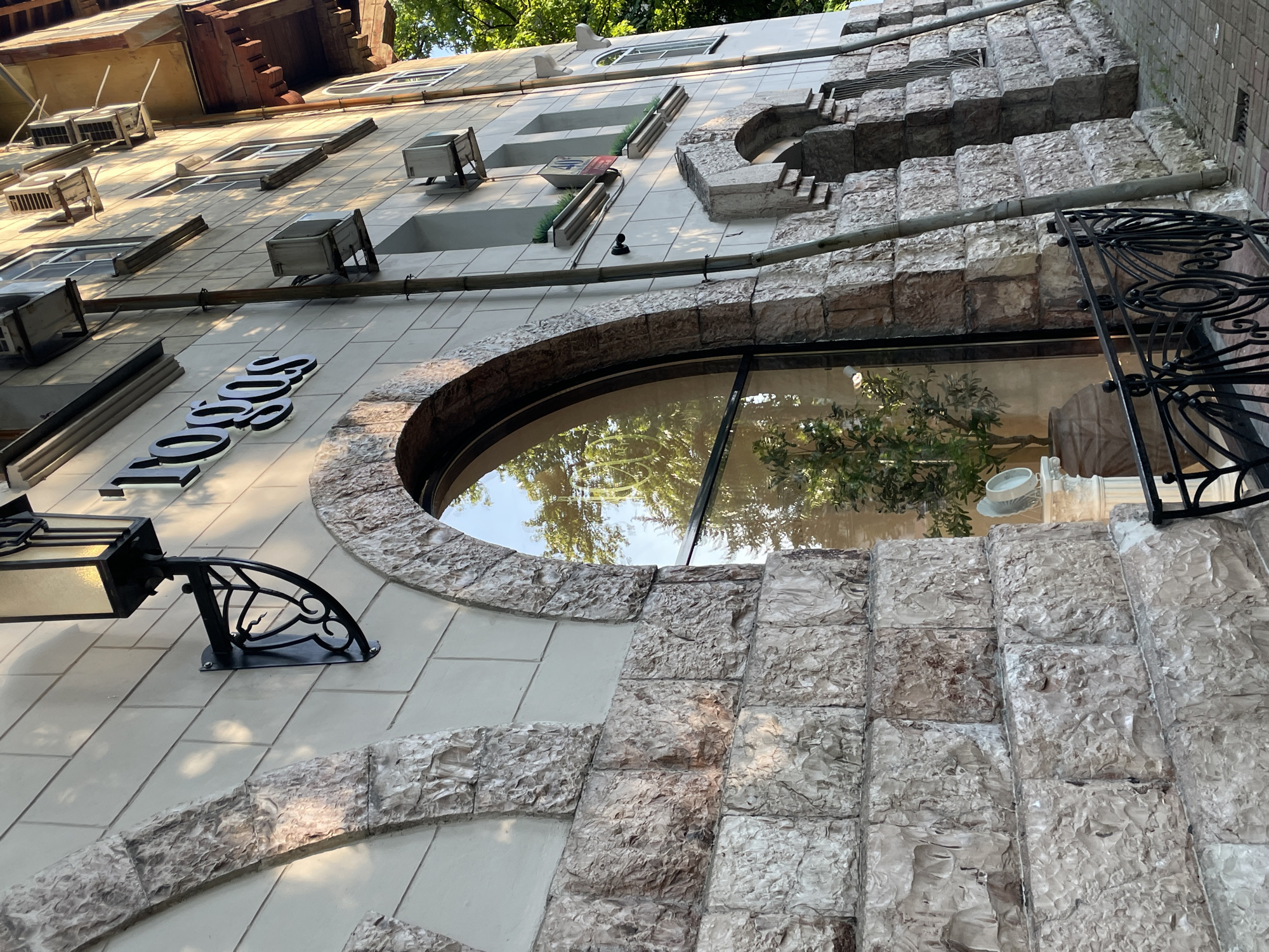 The image shows a building with stone and tiled facade, located in the city of Sochi, Russia. The building features an arched window with a reflection of trees and sky, and a sign that reads "Logos". There are air conditioning units mounted on the facade, as well as an outdoor lamp. A black metal fence is visible in the foreground. The overall scene depicts the exterior of a commercial building.