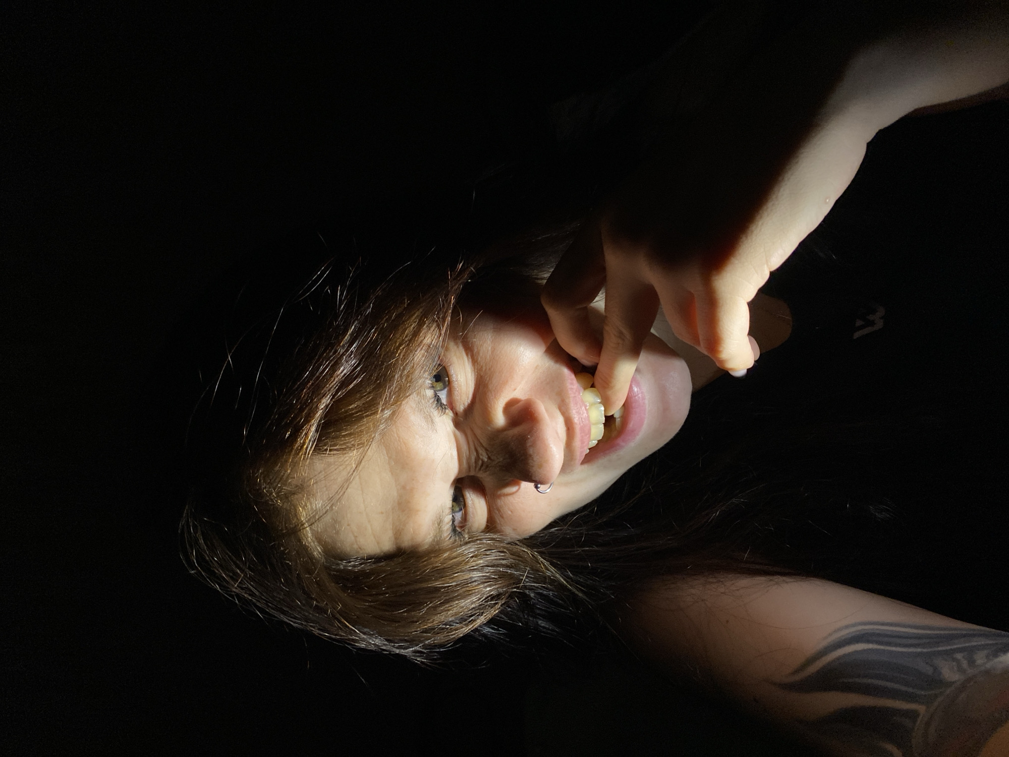 A low-angle, medium shot shows a woman with brown hair and fair skin in a dark setting. Her right hand is raised to her mouth, with a finger touching her teeth. Her eyes are looking directly at the camera, and her brow is furrowed, giving her a menacing expression. She is wearing a dark top, and a tattoo is visible on her left arm. The lighting creates harsh shadows on her face, emphasizing the dramatic expression. The background is entirely black. There is no indication of Chaikovsky, Russia, in the image.