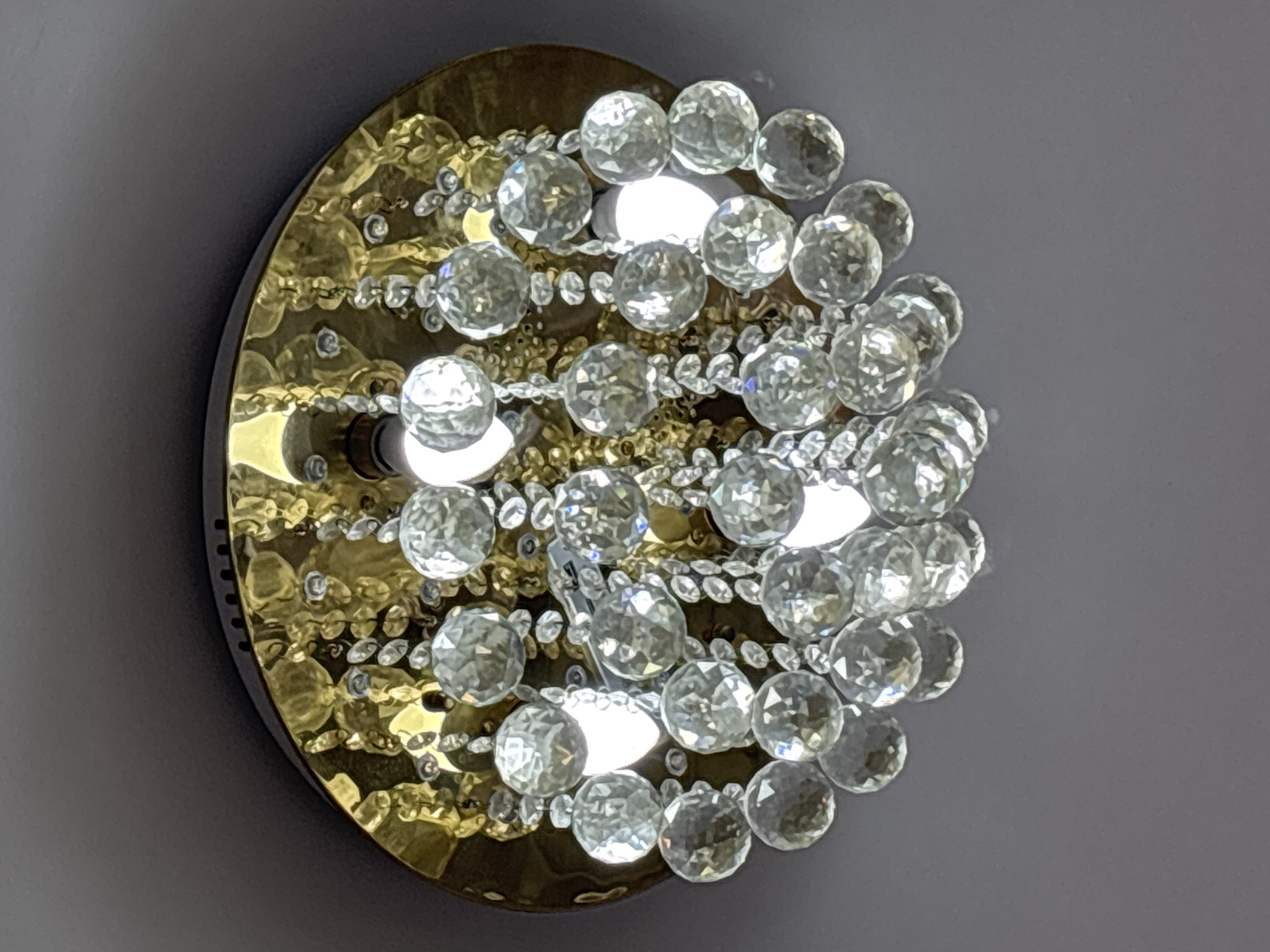 This image captures a close-up view of a circular, ornate light fixture, likely a wall sconce, mounted against a plain, light gray wall. The central subject is the fixture itself, which features a prominent golden, metallic base. This base has a distinct, textured surface, appearing hammered or rippled, creating a rich, reflective pattern.
Adorning the golden base are numerous small, clear, faceted crystal beads or balls. These crystals are arranged in dense clusters and strands, reflecting light brightly and creating a shimmering effect. Several internal light sources are visible as bright, white circular glows emanating from within the fixture, illuminating the crystals. These are likely small LED bulbs or similar light elements integrated into the design.
No people are present, and no activity or event is depicted. The image is a static shot focusing on the decorative object. The background is uniformly light gray, serving as a neutral backdrop that highlights the intricate details and reflective qualities of the fixture. The design suggests a piece of decorative lighting intended for an indoor setting. No text is visible in the image. The provided location context is Sofia, Bulgaria, though this is not visually discernible from the image itself.