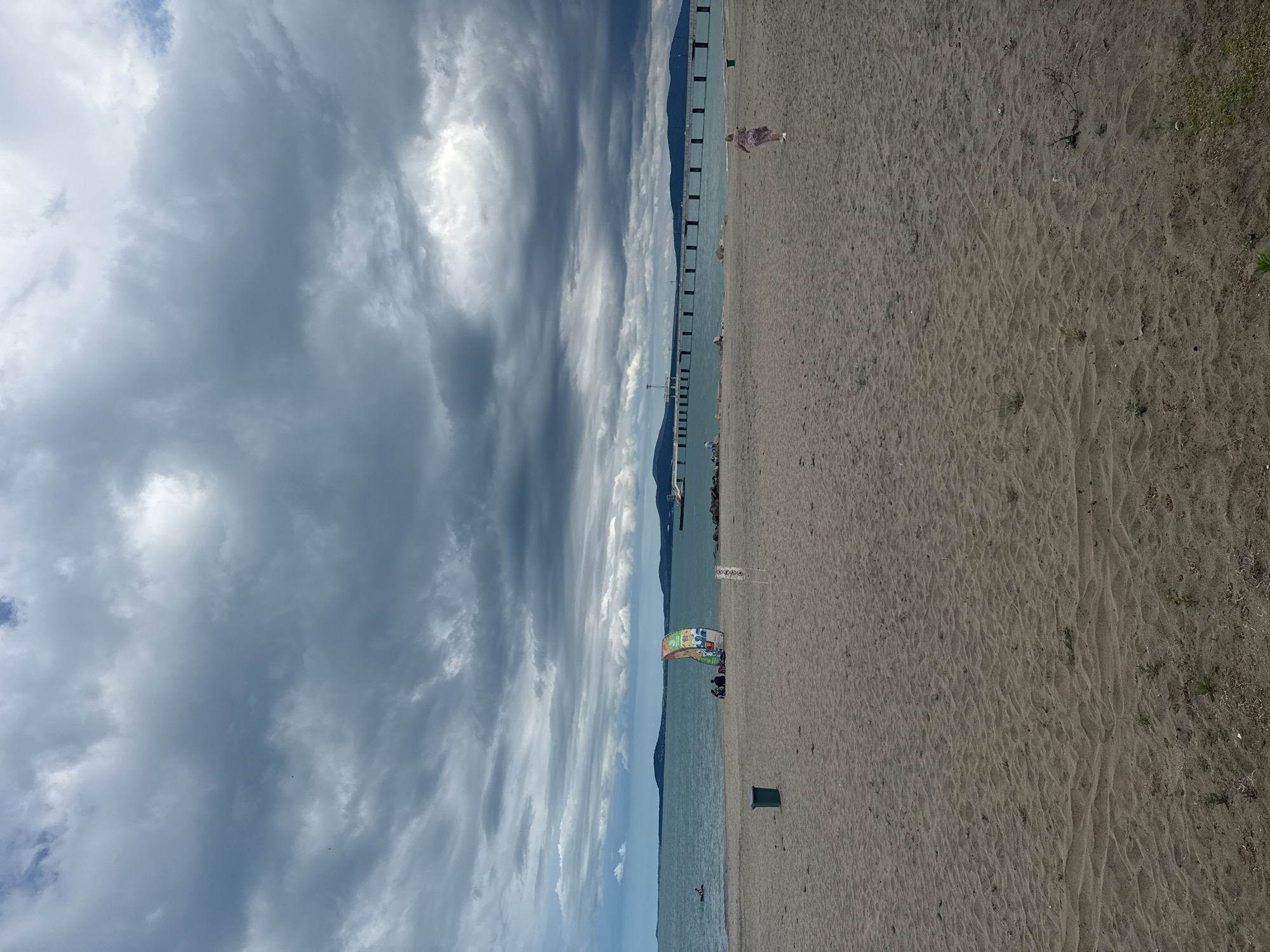 This is an image of the Burgas beach in Burgas, Bulgaria. The scene is a wide sandy beach under a cloudy sky. The sea is calm and a long pier extends into it from the distance. In the mid-ground, a colorful sailboard is positioned on the sand near a few people sitting together. Further to the right, a young girl in a light-colored dress is running towards the right edge of the frame. A dark green trash bin is visible near the water's edge on the left side of the beach. The overall atmosphere is overcast, suggesting a potential change in weather.