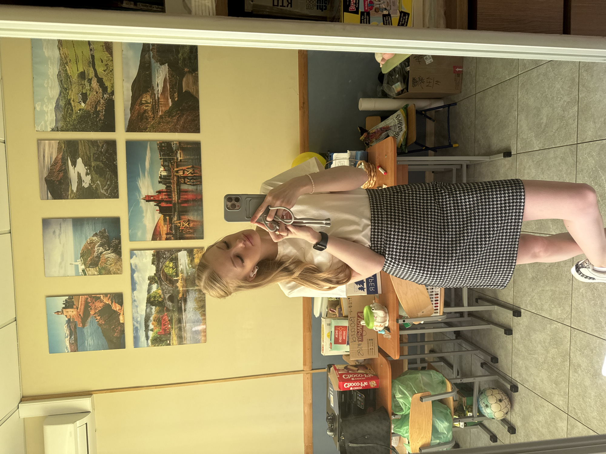 A young woman with blonde hair, wearing a white t-shirt and a black and white houndstooth skirt, takes a selfie in a mirror. She is holding a phone in her left hand and what appears to be a pair of scissors in her right. Her eyes are closed, and she has a slight smile. She is wearing earbuds and a black watch on her left wrist.
The background shows a room with a pale yellow wall adorned with several landscape photographs. To the right, there are shelves filled with various items, including boxes, a yellow balloon, and green plastic bags. A rolling chair and a small desk are also visible. The floor is tiled with a light grey and white pattern. The overall impression is that of a casual indoor setting, possibly a classroom or a studio.
The text visible in the image appears to be in Russian and includes brand names and potentially instructions or advertisements. Some of the legible text fragments are "chi.ru", "Bra", "VANVINE", "CELOTTH", and "Способс". The time of day and weather are not discernible from the image. The woman's expression suggests a relaxed and playful mood.