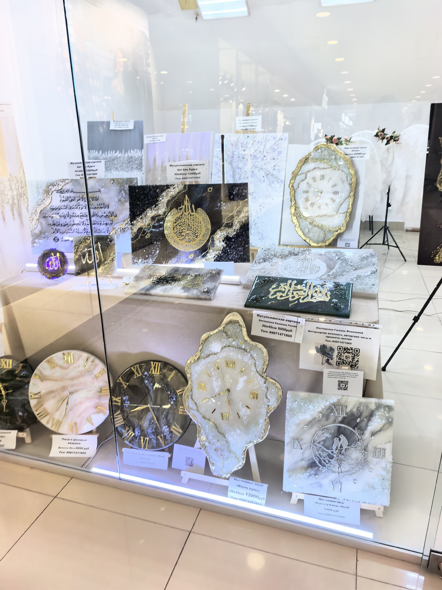 This image shows a display of handmade resin art and clocks, likely in a shop or market stall in Sterlitamak, Russia. The display is behind a glass window, suggesting it's a shopfront or a window display. The items include several large circular clocks with Roman numerals and intricate designs, as well as framed pieces of art, some of which appear to be religious calligraphy. One prominent piece of art features Arabic script in gold on a dark background with sparkling accents. Other artworks have a marbled effect with gold detailing. Prices and contact information, including phone numbers and QR codes, are visible on small white labels placed in front of some of the items. The overall aesthetic is modern and luxurious, with a focus on gold and shimmering materials. The background is brightly lit, with white walls and ceiling visible.