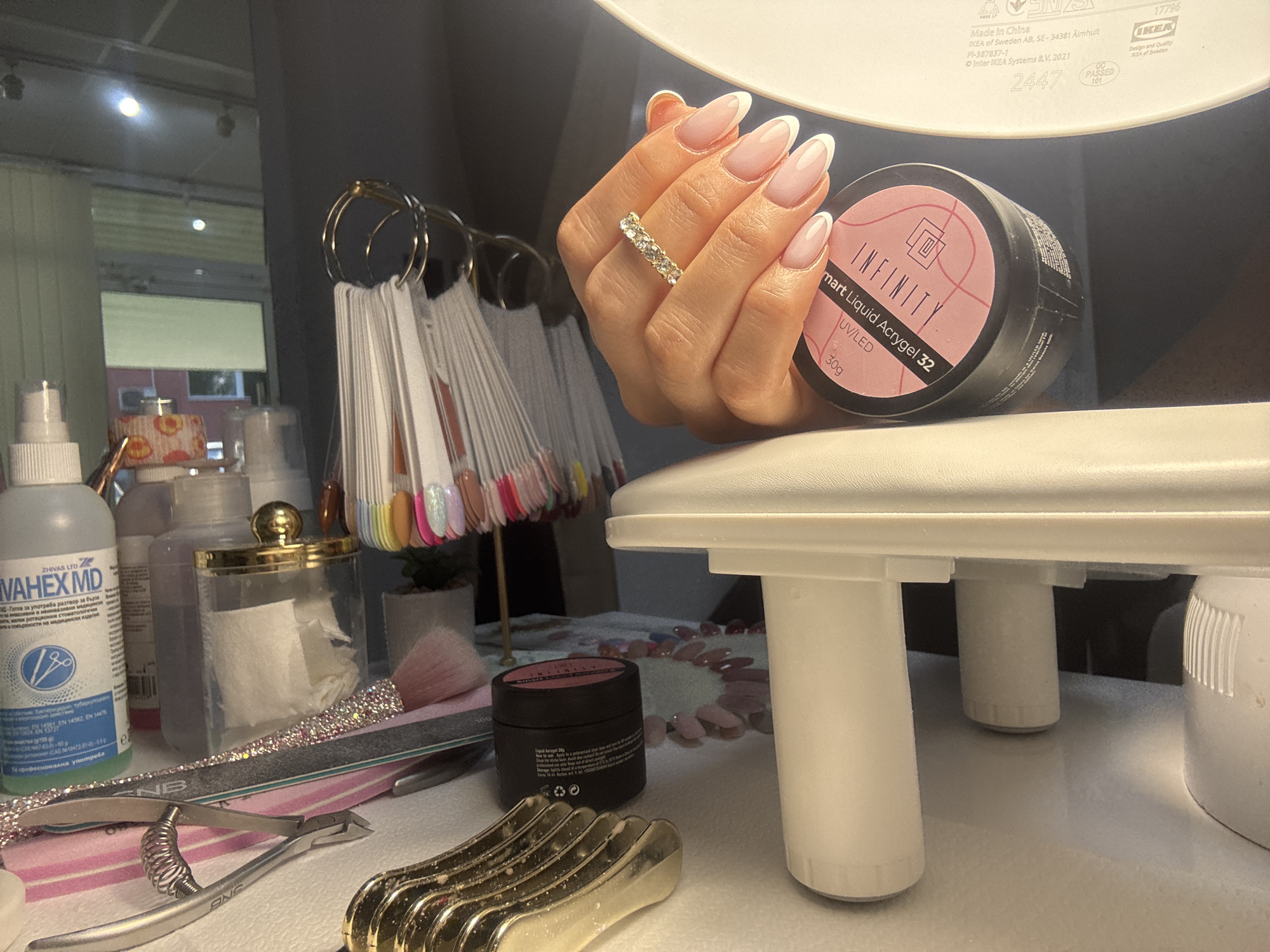This image captures a scene likely within a nail salon setting. The main subject is a hand with a fresh set of French manicure nails, adorned with a diamond ring, holding a container labeled "INFINITY smart Liquid Acrygel 32".
Various objects suggest a nail care environment: a bottle of "VAHEX MD" disinfectant, nail polish color samples on a stand, tools like cuticle nippers and a nail file, cotton pads in a jar, and other nail care products. A white lamp dominates the upper section of the image, illuminating the scene.
The presence of Cyrillic writing on the "VAHEX MD" bottle suggests an Eastern European location, specifically Bulgarian, based on similar product availability and labeling practices. While it is difficult to definitively confirm the exact location in Burgas, Bulgaria without additional context, the presence of these items suggests a professional nail care service within the city.